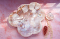 A cloud-shaped pink carnelian flower agate tray with natural stone inclusions, displayed on a pink marbled surface alongside a small clear crystal and dried grass stems.