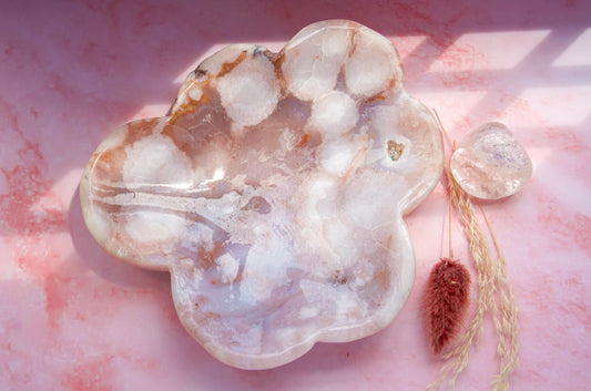 A cloud-shaped pink carnelian flower agate tray with natural stone inclusions, displayed on a pink marbled surface alongside a small clear crystal and dried grass stems.