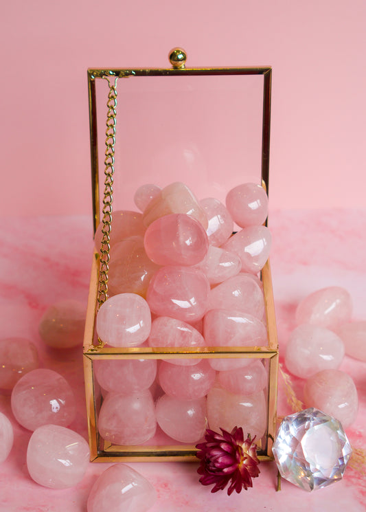 A gold-framed glass box filled with smooth brazilian rose quartz pink tumbled stones, set on a pink surface with more pink stones scattered around and a faceted clear gemstone in front.