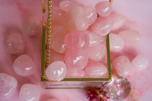 Brazilian rose quartz tumbled stones arranged inside a gold-framed glass box on a soft pink background, with a few stones scattered around and a clear faceted crystal near the bottom right.