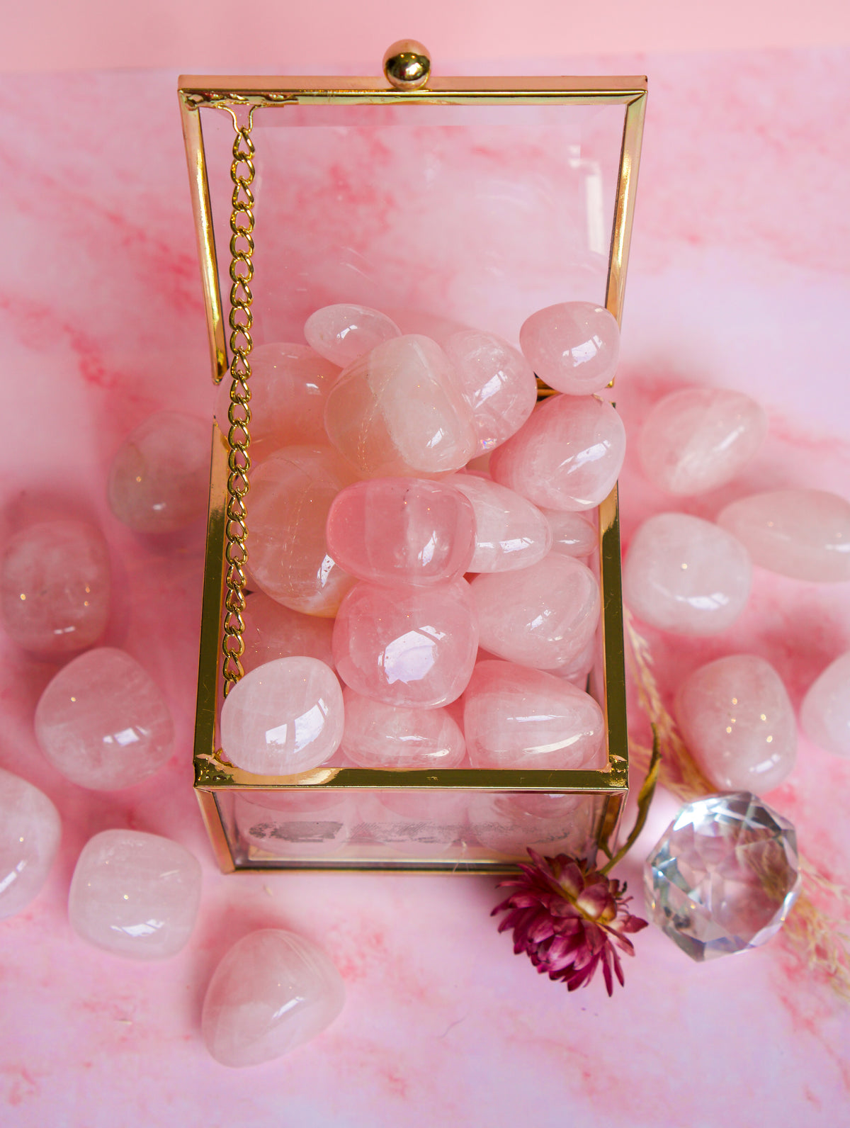 A gold-framed glass box filled with smooth Brazilian rose quartz pink tumbled stones, styled on a pink marble surface with additional pink stones and a small crystal ornament around it.