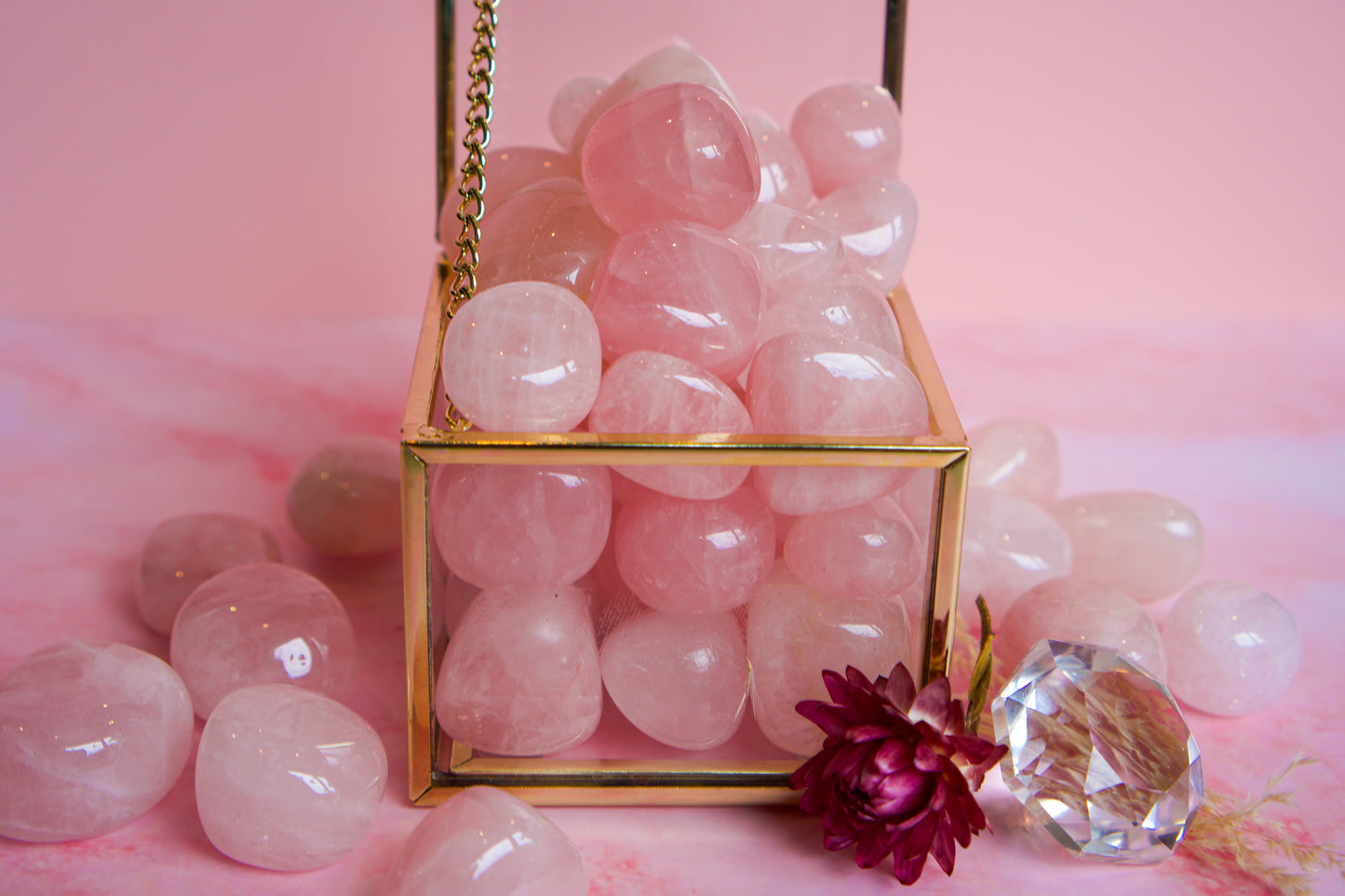 A gold-framed glass box filled with smooth, polished pink Brazilian rose quartz stones, styled on a pink surface with additional loose pink stones and a decorative crystal and flower prop.