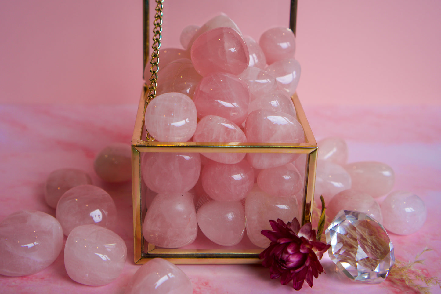 A gold-framed glass cube filled with smooth pink Brazilian rose quartz stones, set on a pink surface with additional loose pink stones and a faceted clear crystal around it.