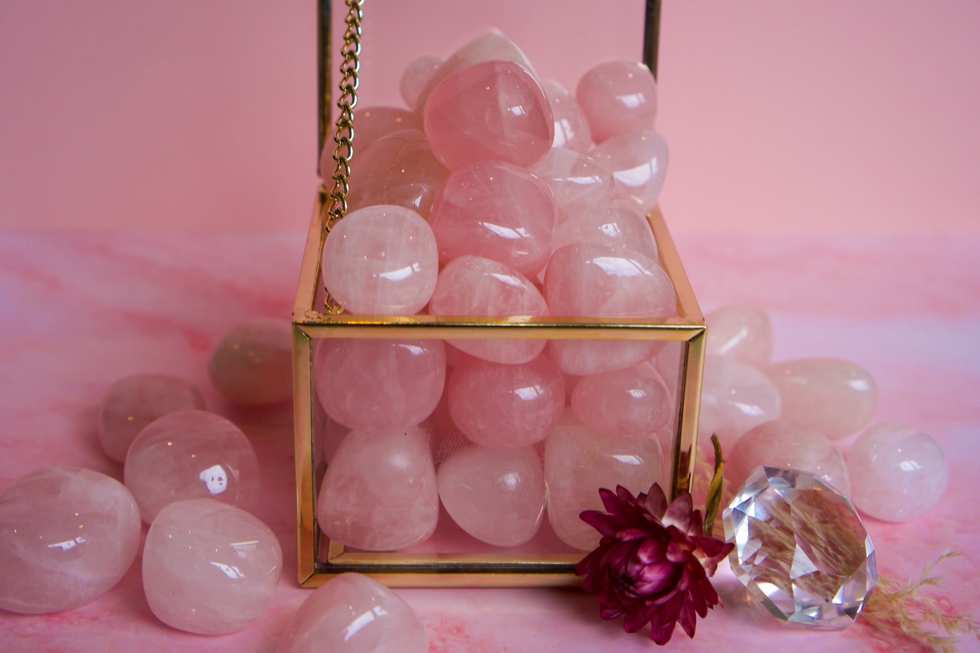 A gold-framed glass cube filled with smooth pink Brazilian rose quartz stones, set on a pink surface with additional loose pink stones and a faceted clear crystal around it.