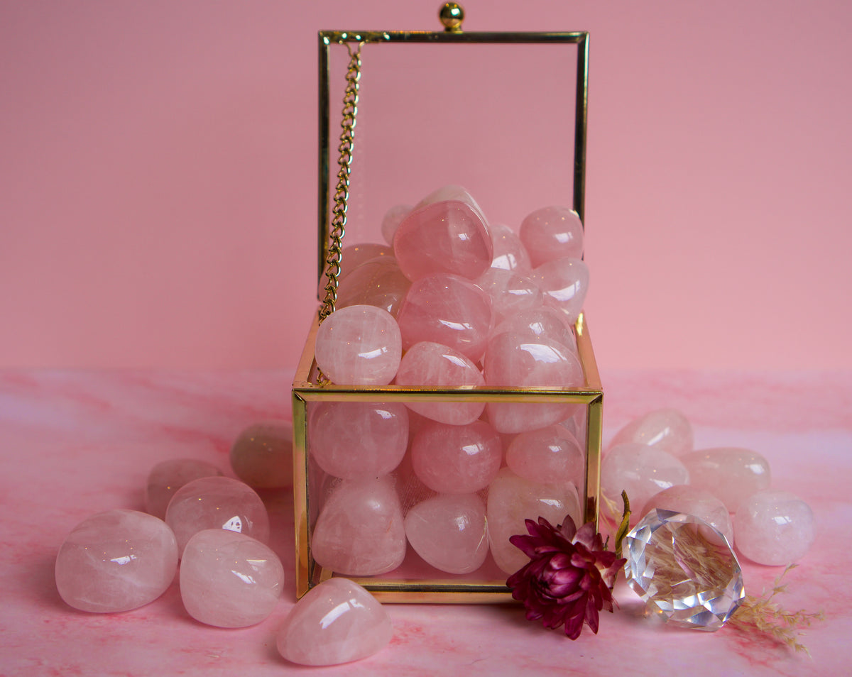 A gold-framed transparent box filled with smooth pink Brazilian rose quartz stones, styled on a pink surface with a few loose stones and a small decorative flower in front.