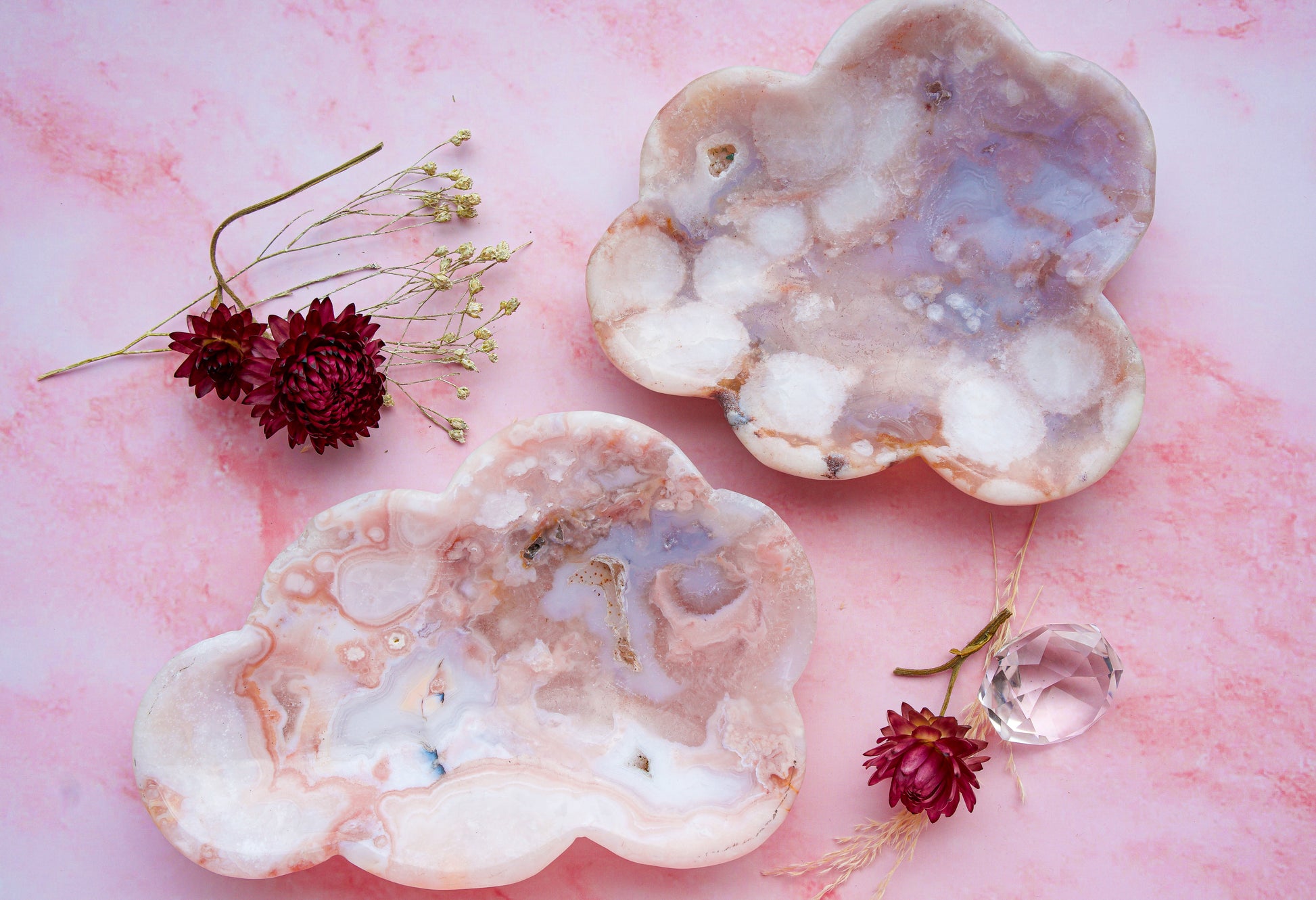 Two cloud-shaped pink carnelian flower agate stone trays with swirling pink, white, and lavender geode patterns, styled on a pink marble background with dried flowers and a clear crystal.