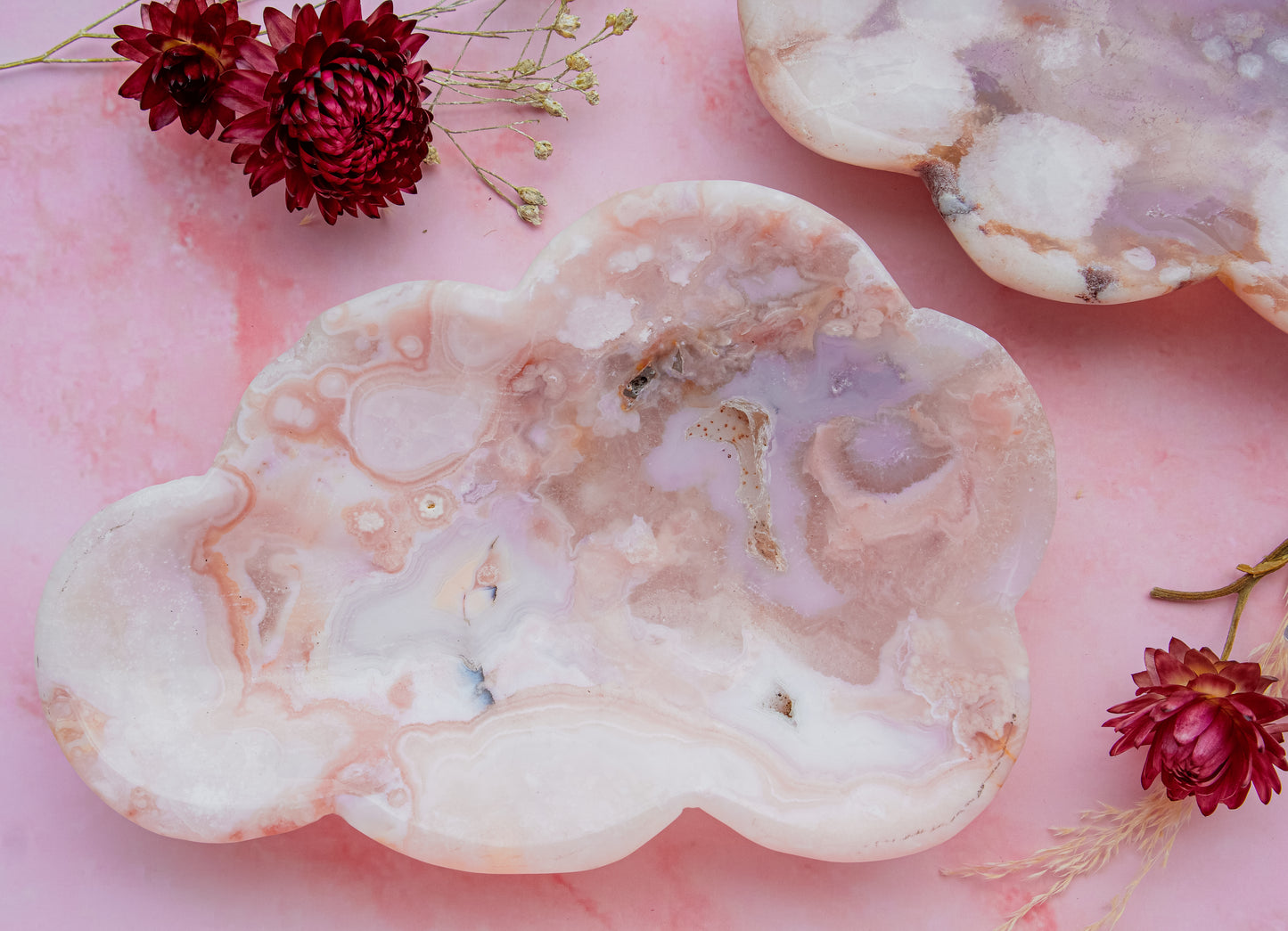 A cloud-shaped pink carnelian flower agate tray carved from pink and blue with purple veining, displayed on a pink background with dried red flowers.