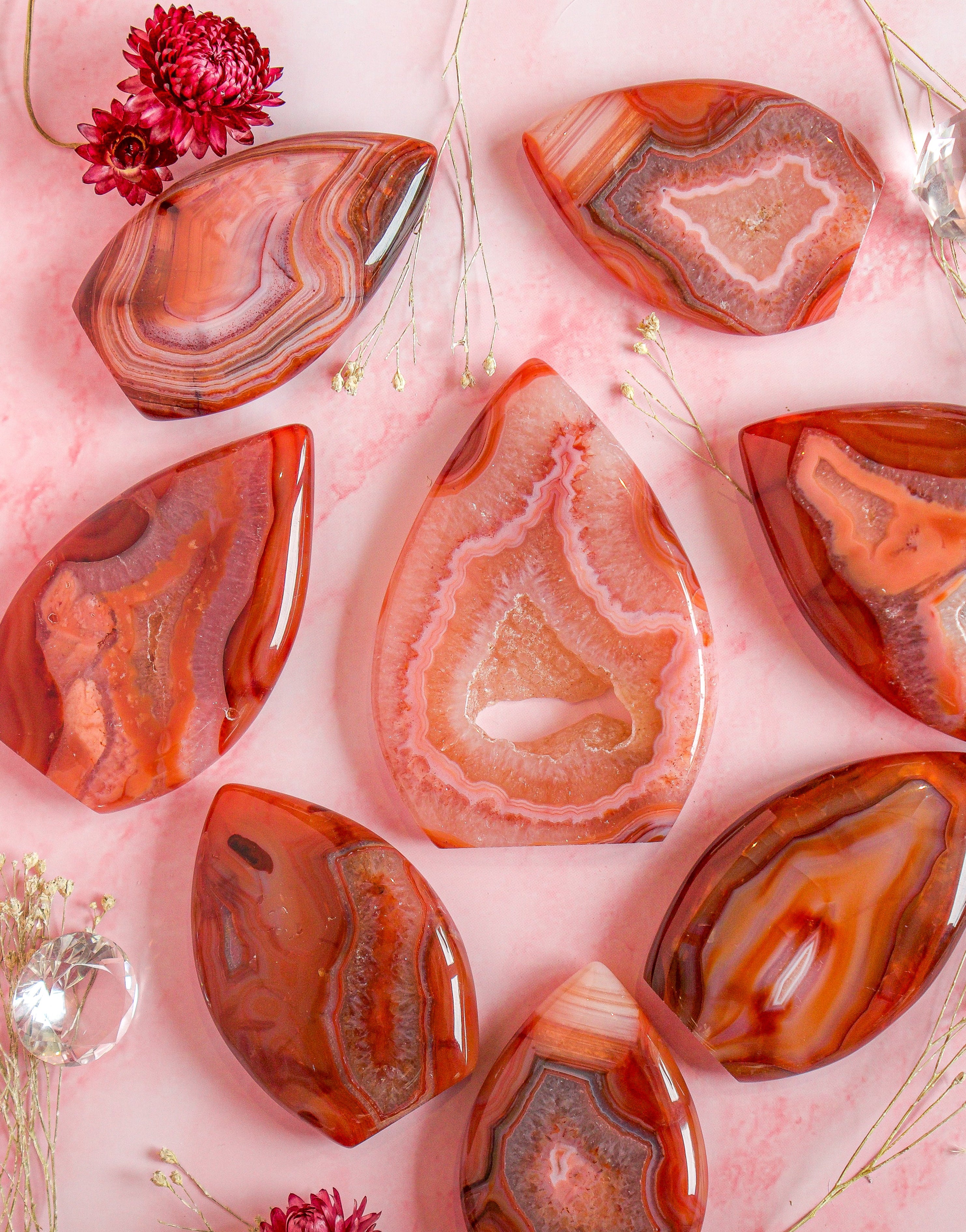 Seven polished red carnelian pink agate stone flame freeforms with natural banding patterns are arranged in a flat-lay on a pink background with dried flowers and crystal spheres.