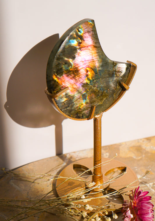 A polished sunset flash rainbow labradorite moon crystal mirror with a gold-toned metal stand, displayed on a marble surface with dried flowers and a warm wall shadow.