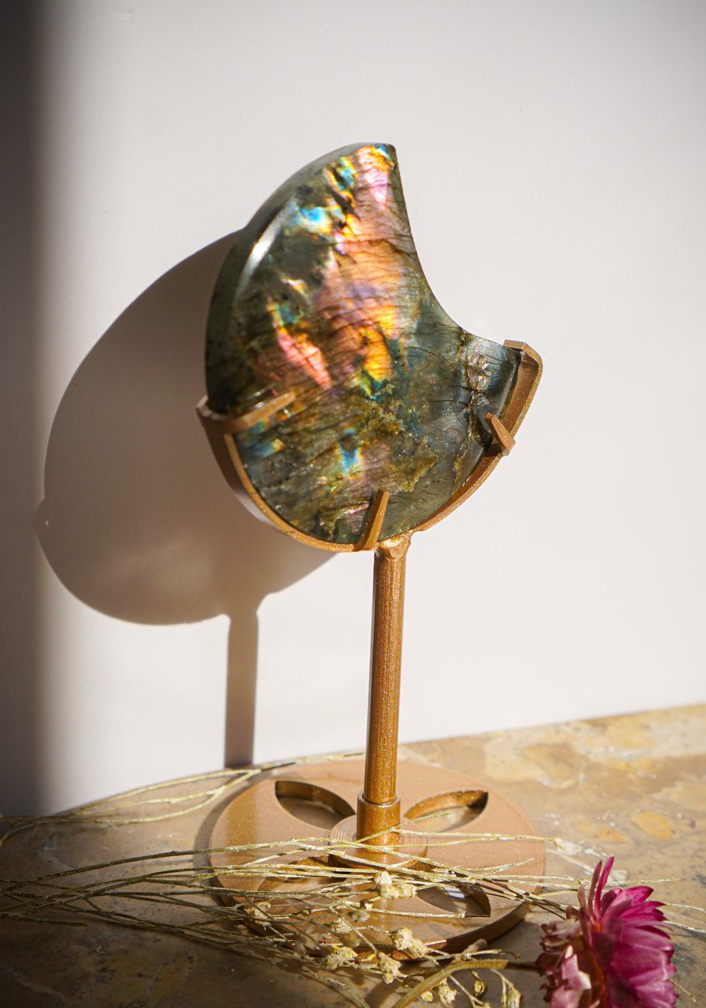 A polished iridescent sunset flash rainbow labradorite moon piece mounted on a gold-toned metal stand, displayed on a marble surface with dried grass and a pink flower against a white wall.