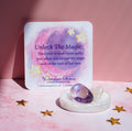 A square card with a pastel moon and star illustration reads 'Unlock The Magic' and includes the brand name Wonder Gems. In front of the card is a white ceramic dish holding a purple amethyst and a clear crystal, set on a pink surface with scattered gold star confetti.