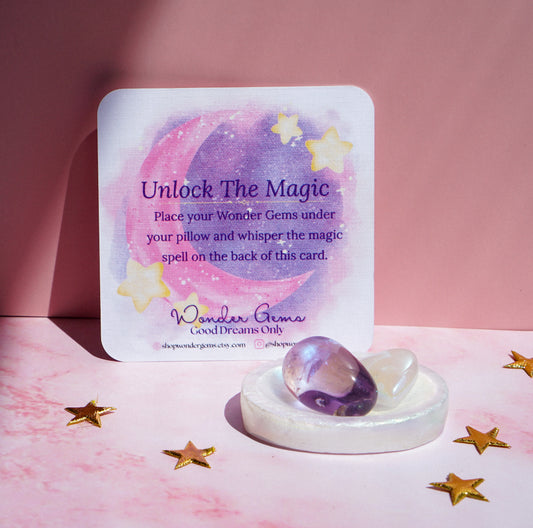 A square card with a pastel moon and star illustration reads 'Unlock The Magic' and includes the brand name Wonder Gems. In front of the card is a white ceramic dish holding a purple amethyst and a clear crystal, set on a pink surface with scattered gold star confetti.