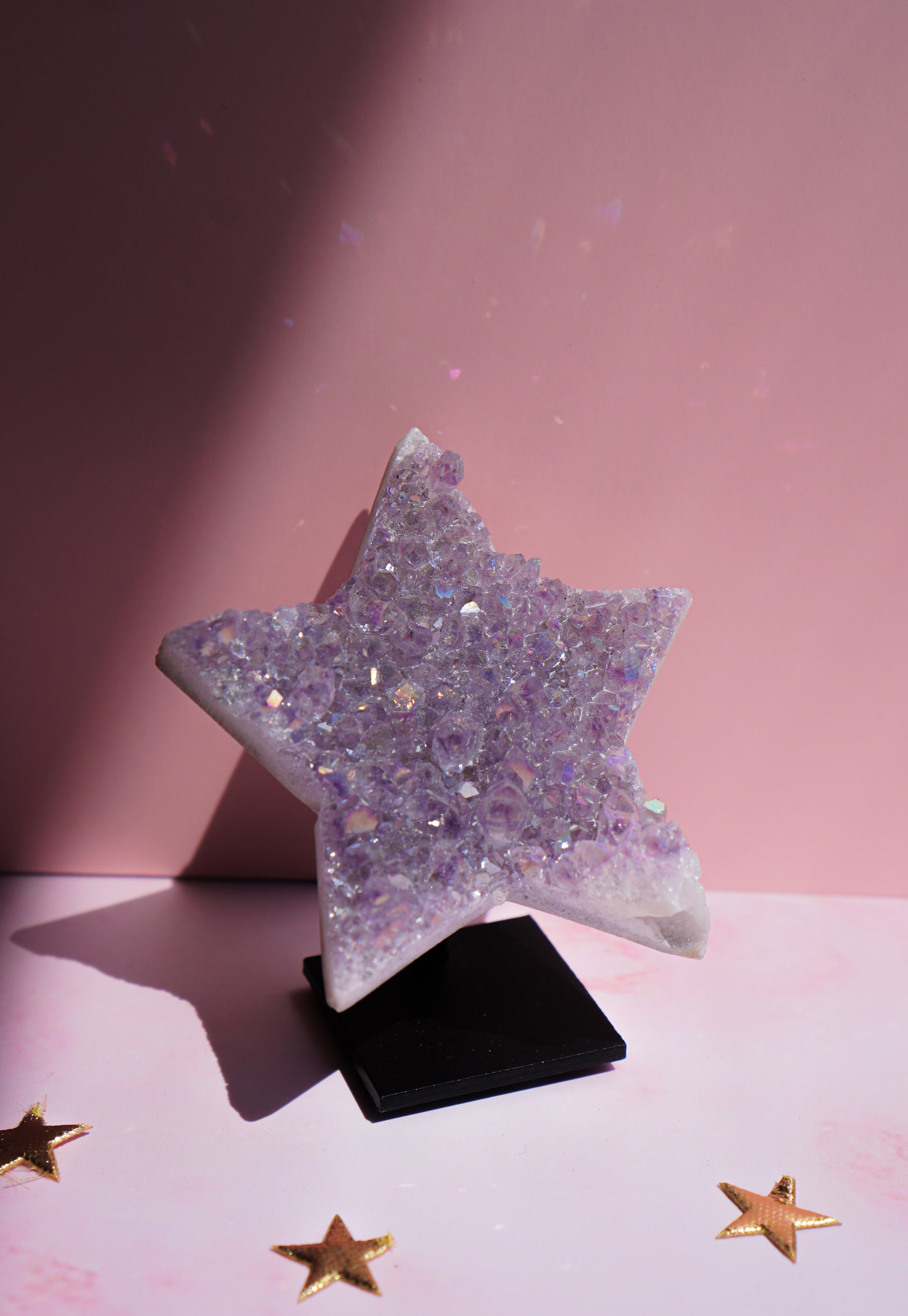 A five-pointed star-shaped Angel aura amethyst crystal raw carving cluster with iridescent lavender and white faceted stones, displayed on a black stand against a pink background with decorative gold stars on the surface.