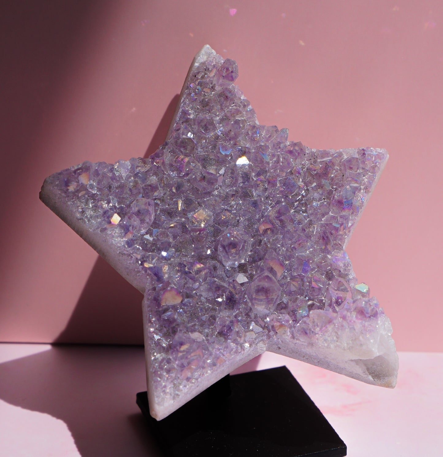 A five-pointed star-shaped Angel aura amethyst crystal raw carving cluster display made of iridescent lavender and purple faceted stones, mounted on a black display stand against a pink background.