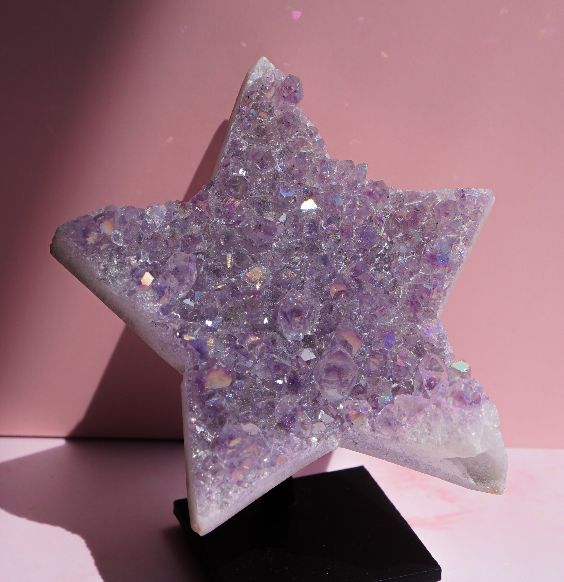 A five-pointed star-shaped Angel aura amethyst crystal raw carving cluster display made of iridescent lavender and purple faceted stones, mounted on a black display stand against a pink background.