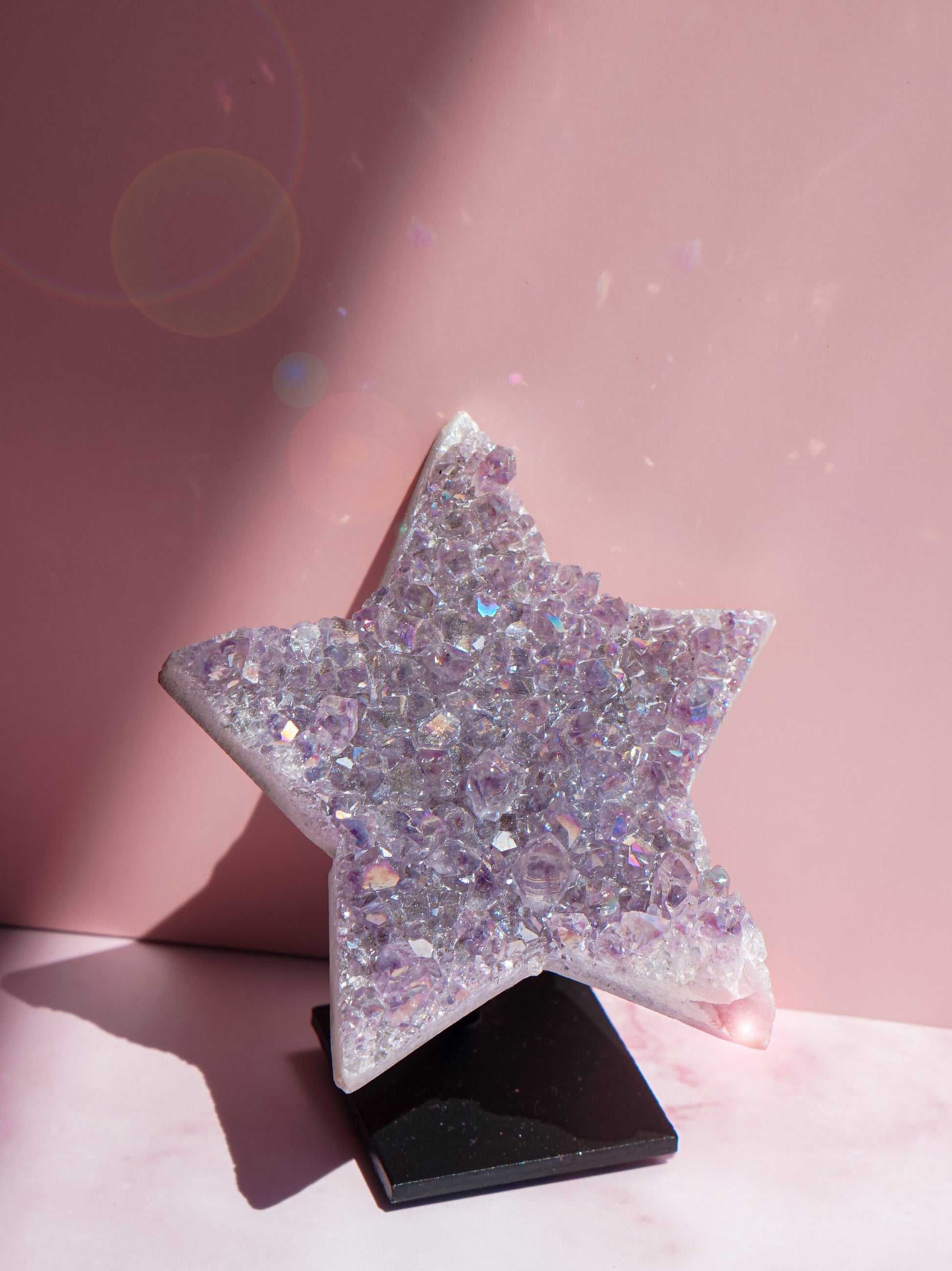 A five-pointed star-shaped Aura Amethyst raw crystal carved cluster standee covered in iridescent lavender and silver rhinestones, displayed on a small black base against a pink background.