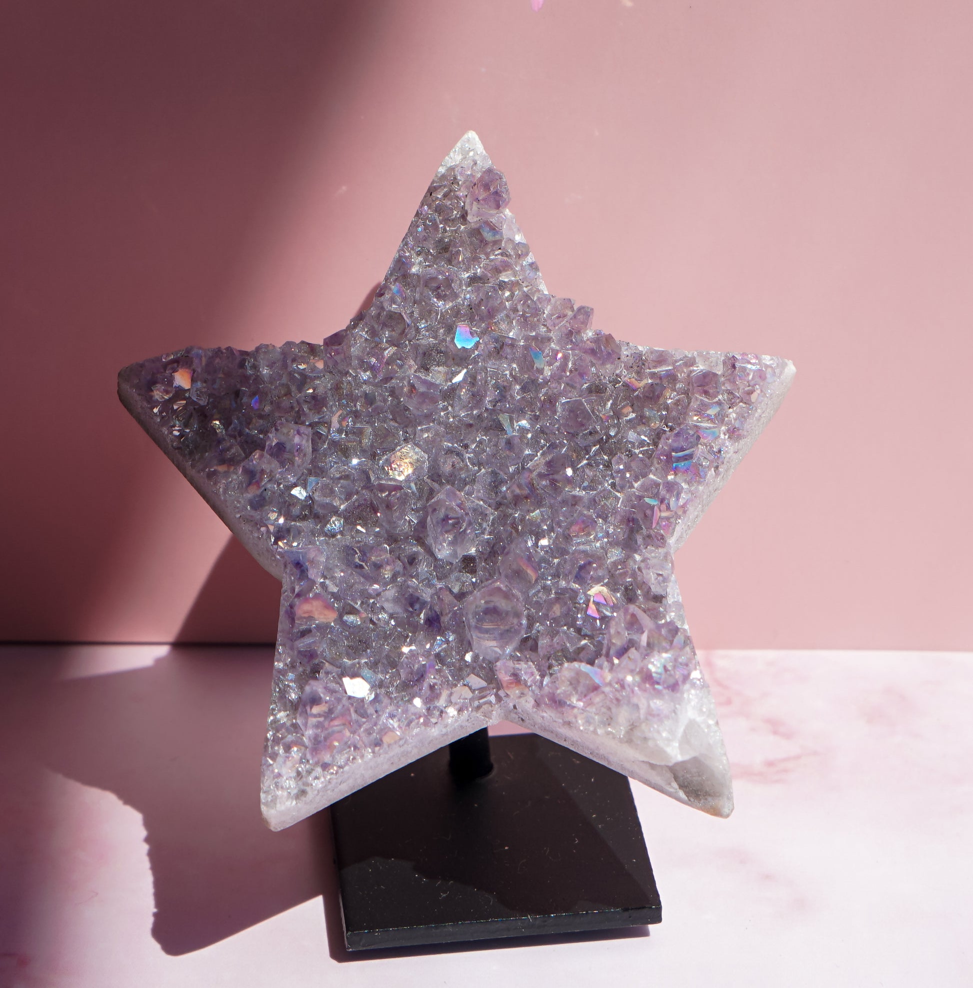A five-pointed star-shaped Aura Amethyst raw crystal carved cluster with iridescent lavender and purple hues, displayed on a black stand against a pink background.