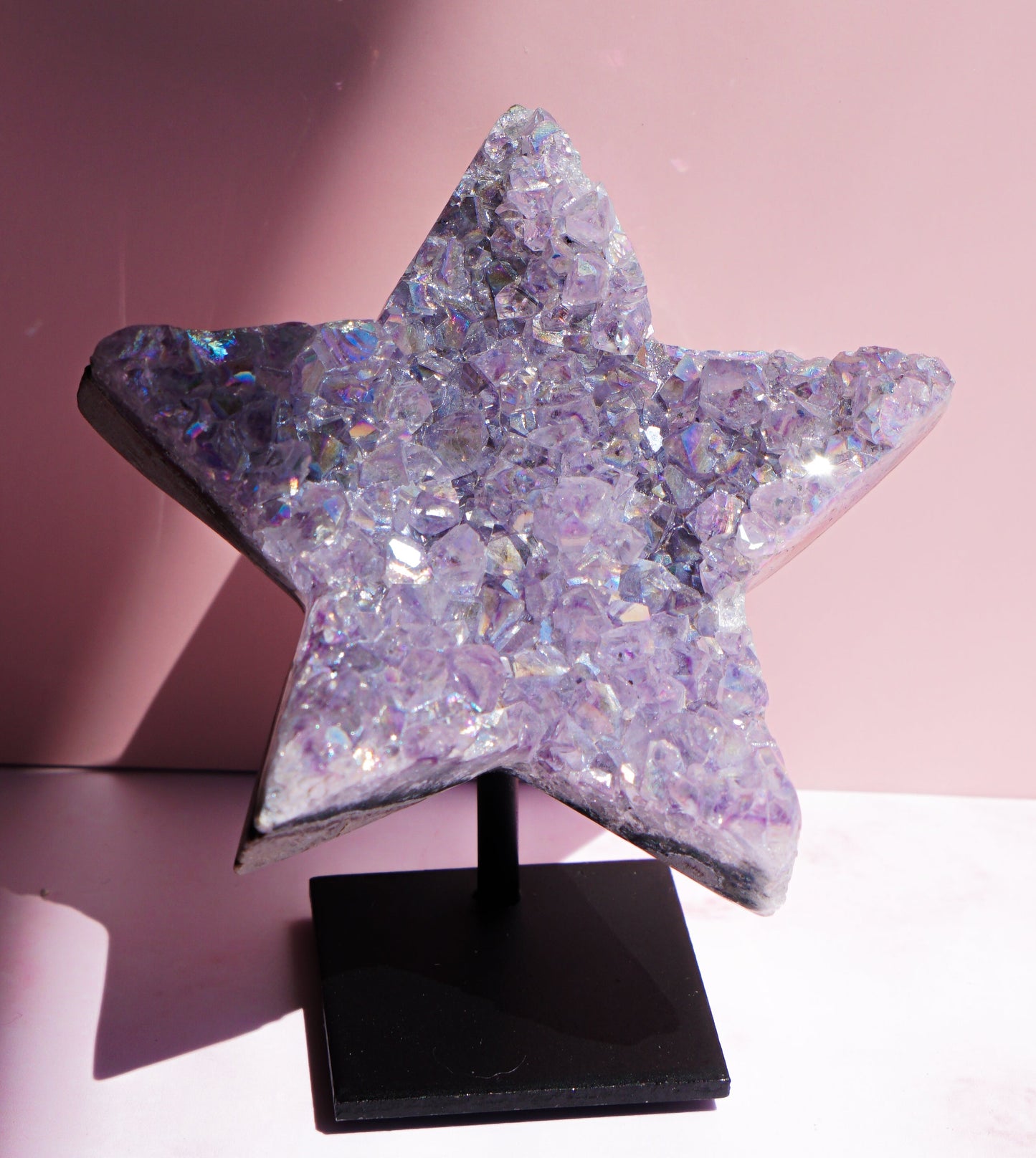 A five-pointed star-shaped Aura Amethyst raw crystal carved cluster with iridescent lavender and purple hues, displayed on a black stand against a pink background.