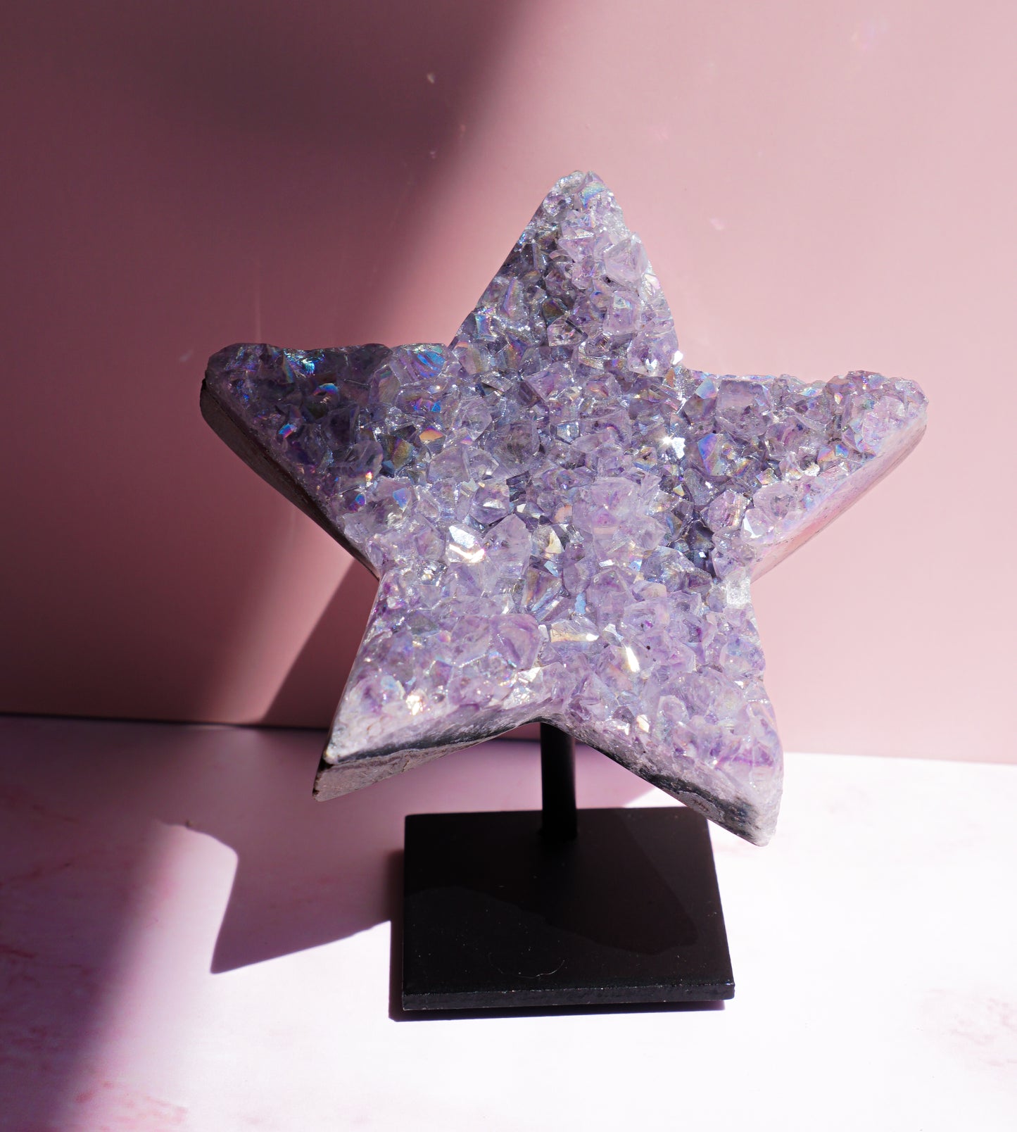 A five-pointed star-shaped Aura Amethyst raw crystal carved cluster with iridescent lavender and purple hues, displayed on a black stand against a pink background.