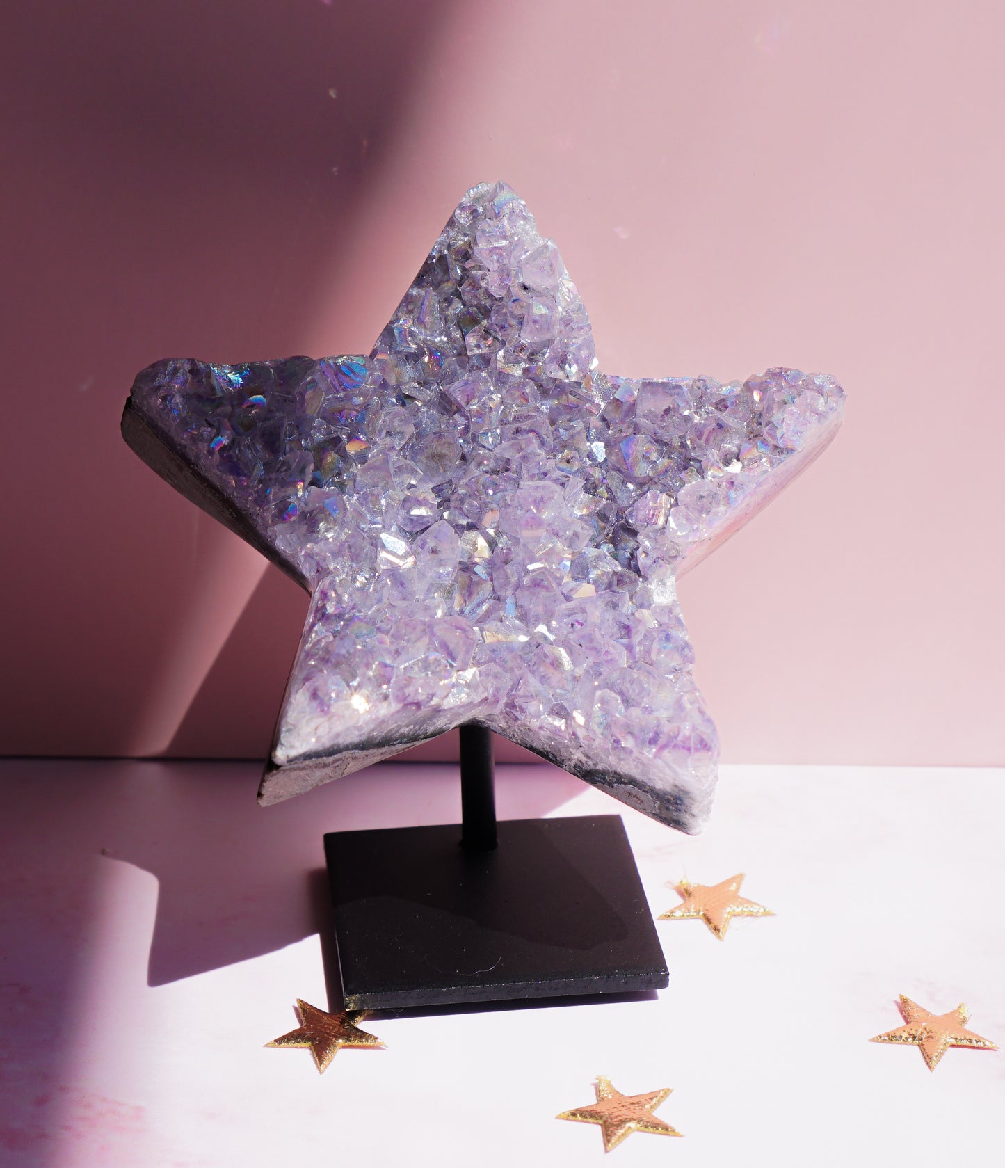 A five-pointed star-shaped Aura Amethyst raw crystal carved cluster with iridescent lavender and purple hues, displayed on a black stand against a pink background.