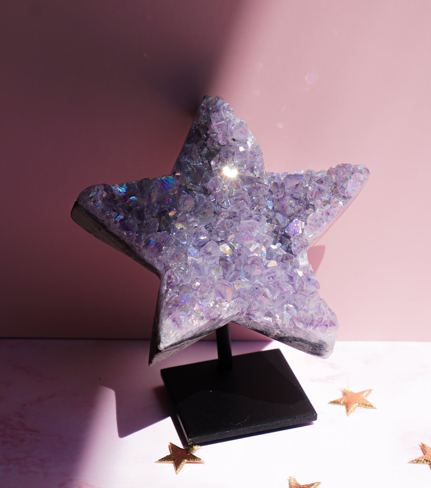A glittery star-shaped decorative object on a black stand, photographed against a pink studio background with a few small gold star confetti pieces on the surface.