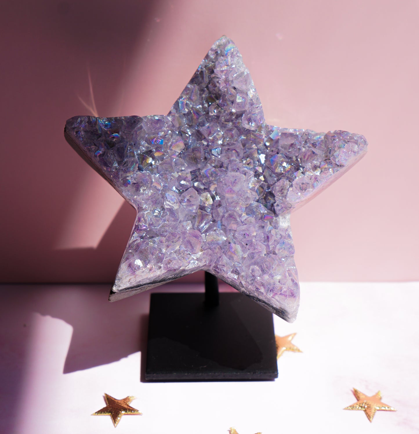 A five-pointed star-shaped Aura Amethyst raw crystal carved cluster with iridescent lavender and purple hues, displayed on a black stand against a pink background.