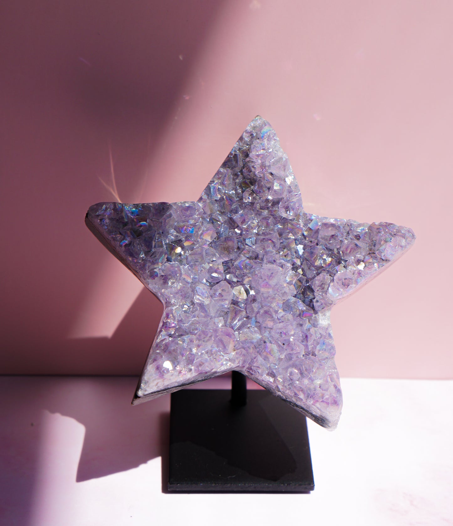 A five-pointed star-shaped Aura Amethyst raw crystal carved cluster with iridescent lavender and purple hues, displayed on a black stand against a pink background.