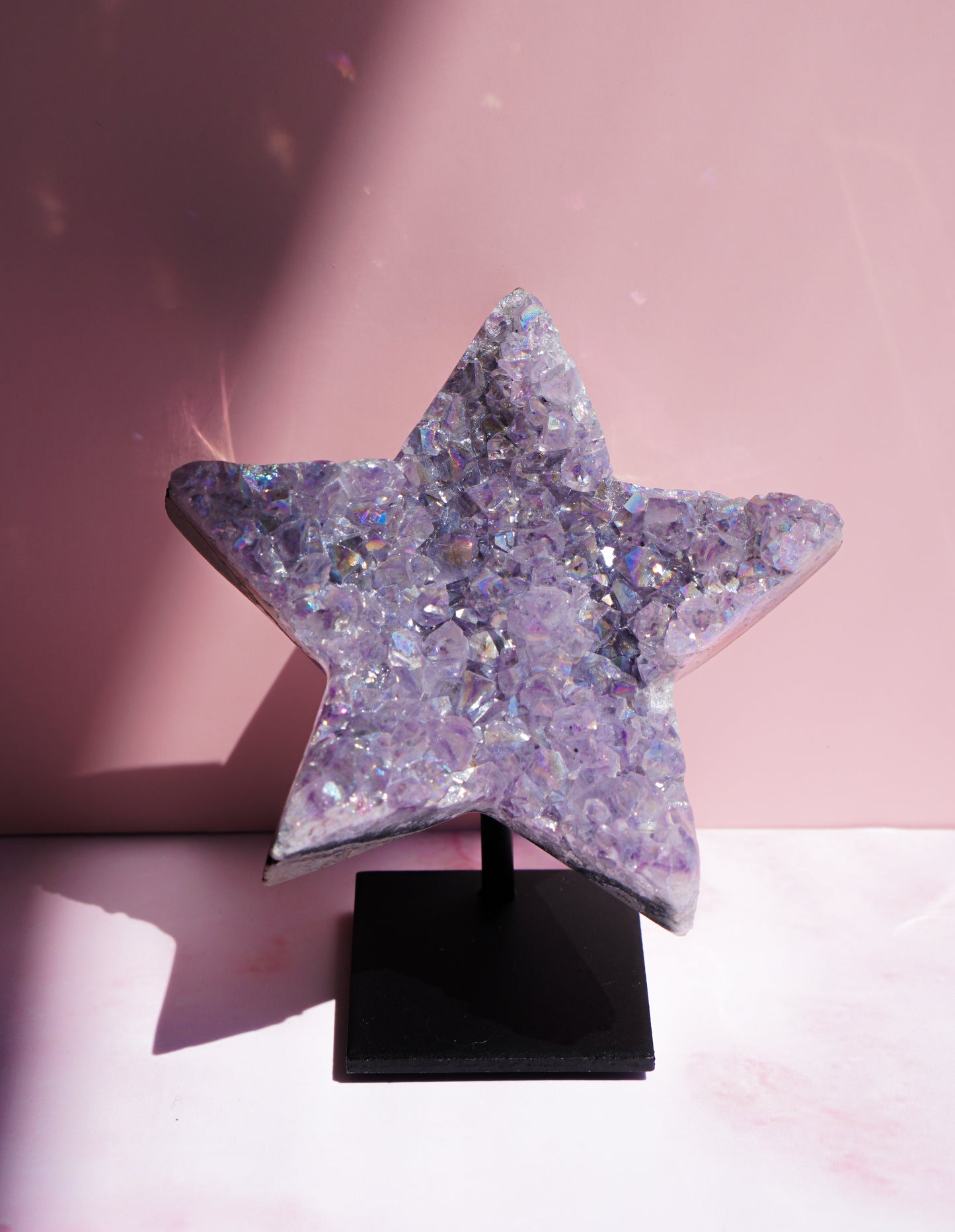 A five-pointed star-shaped Aura Amethyst raw crystal carved cluster with iridescent lavender and purple hues, displayed on a black stand against a pink background.