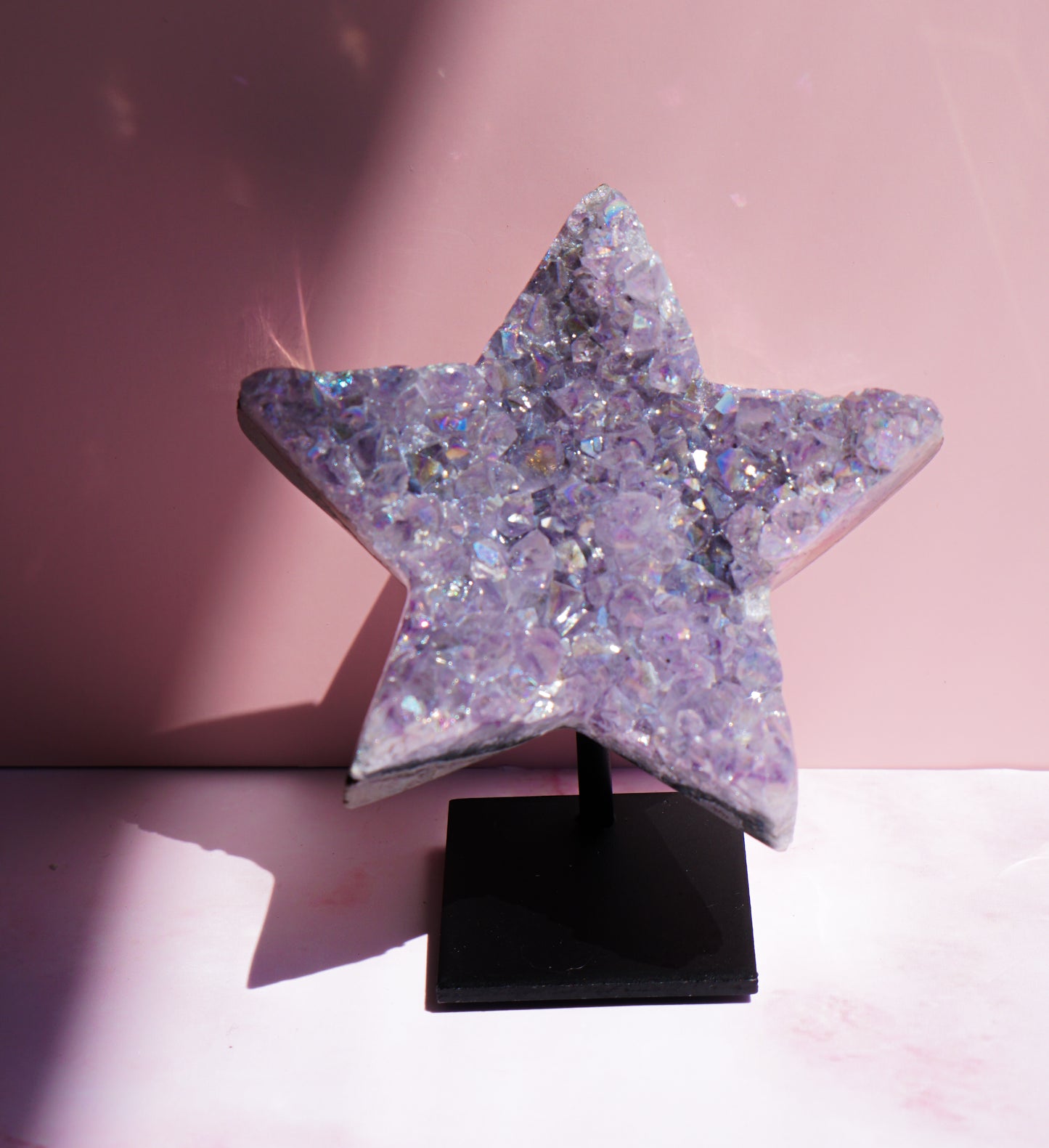 A five-pointed star-shaped Aura Amethyst raw crystal carved cluster with iridescent lavender and purple hues, displayed on a black stand against a pink background.