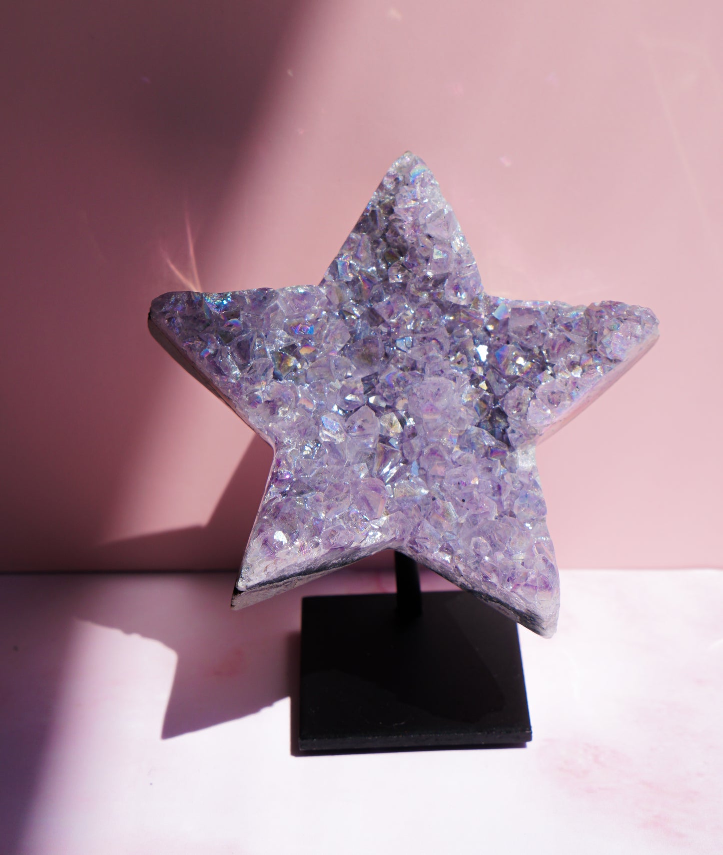 A five-pointed star-shaped Aura Amethyst raw crystal carved cluster with iridescent lavender and purple hues, displayed on a black stand against a pink background.