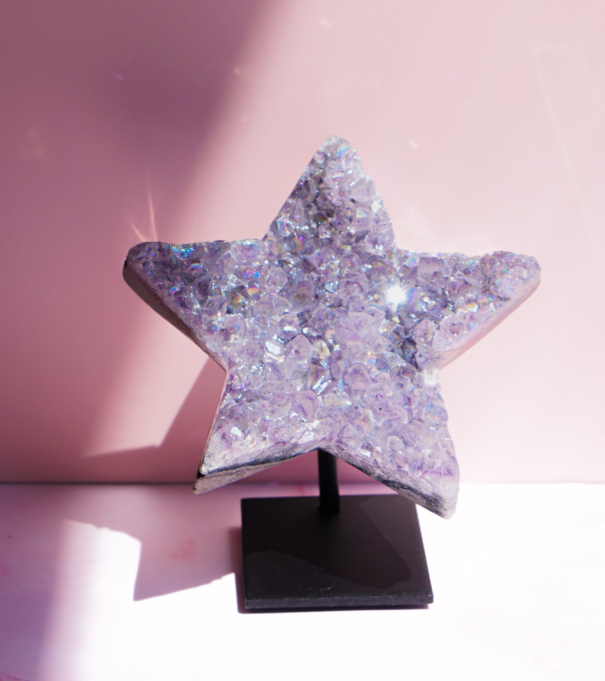 A five-pointed star-shaped Aura Amethyst raw crystal carved cluster with iridescent lavender and purple hues, displayed on a black stand against a pink background.