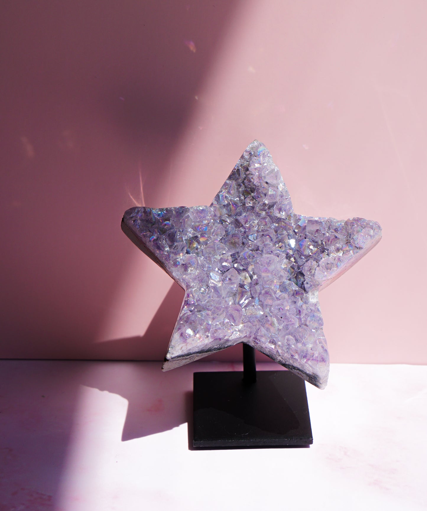 A five-pointed star-shaped Aura Amethyst raw crystal carved cluster with iridescent lavender and purple hues, displayed on a black stand against a pink background.
