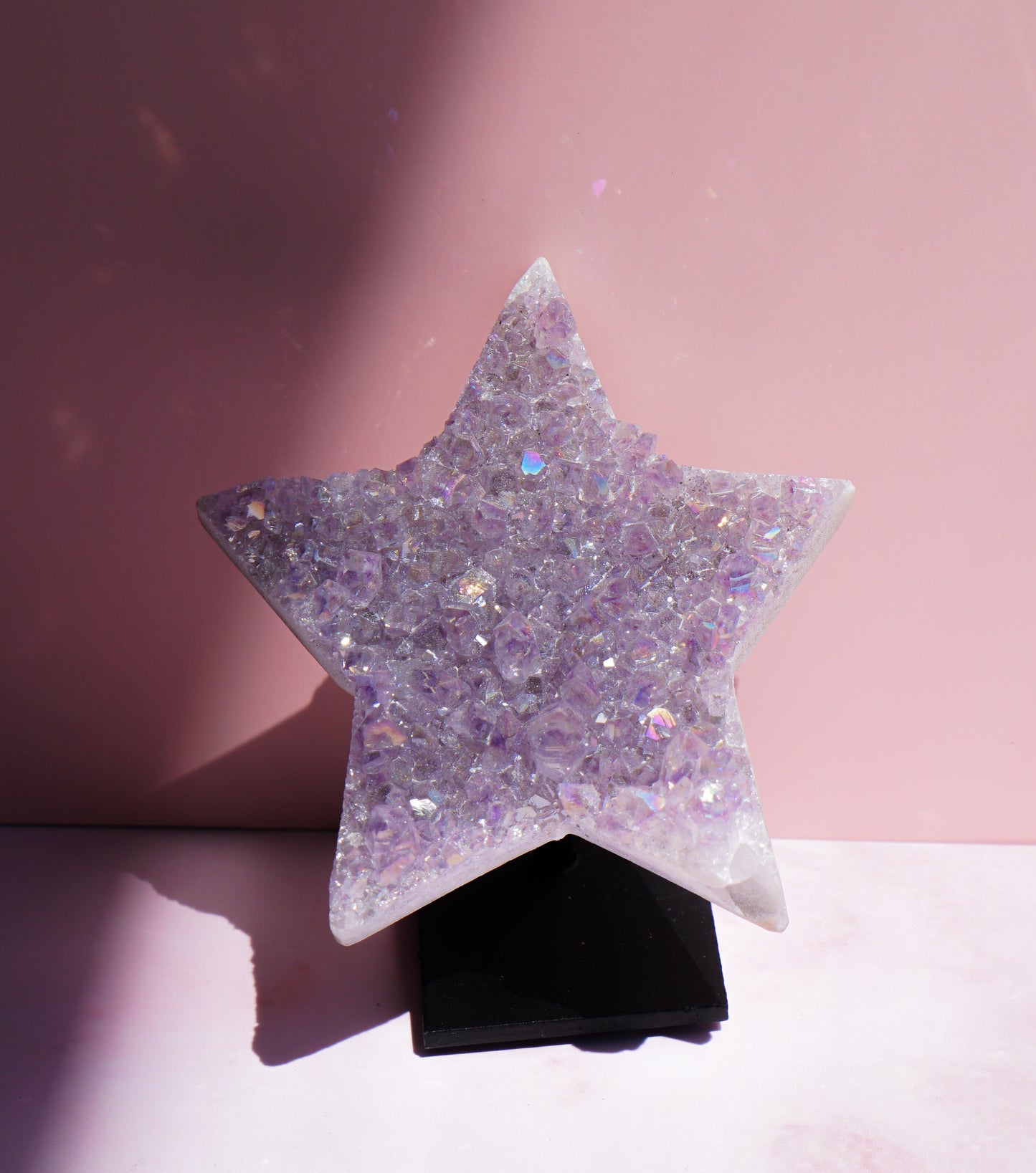 A five-pointed star-shaped Aura Amethyst raw crystal carved cluster with iridescent lavender and purple hues, displayed on a black stand against a pink background.