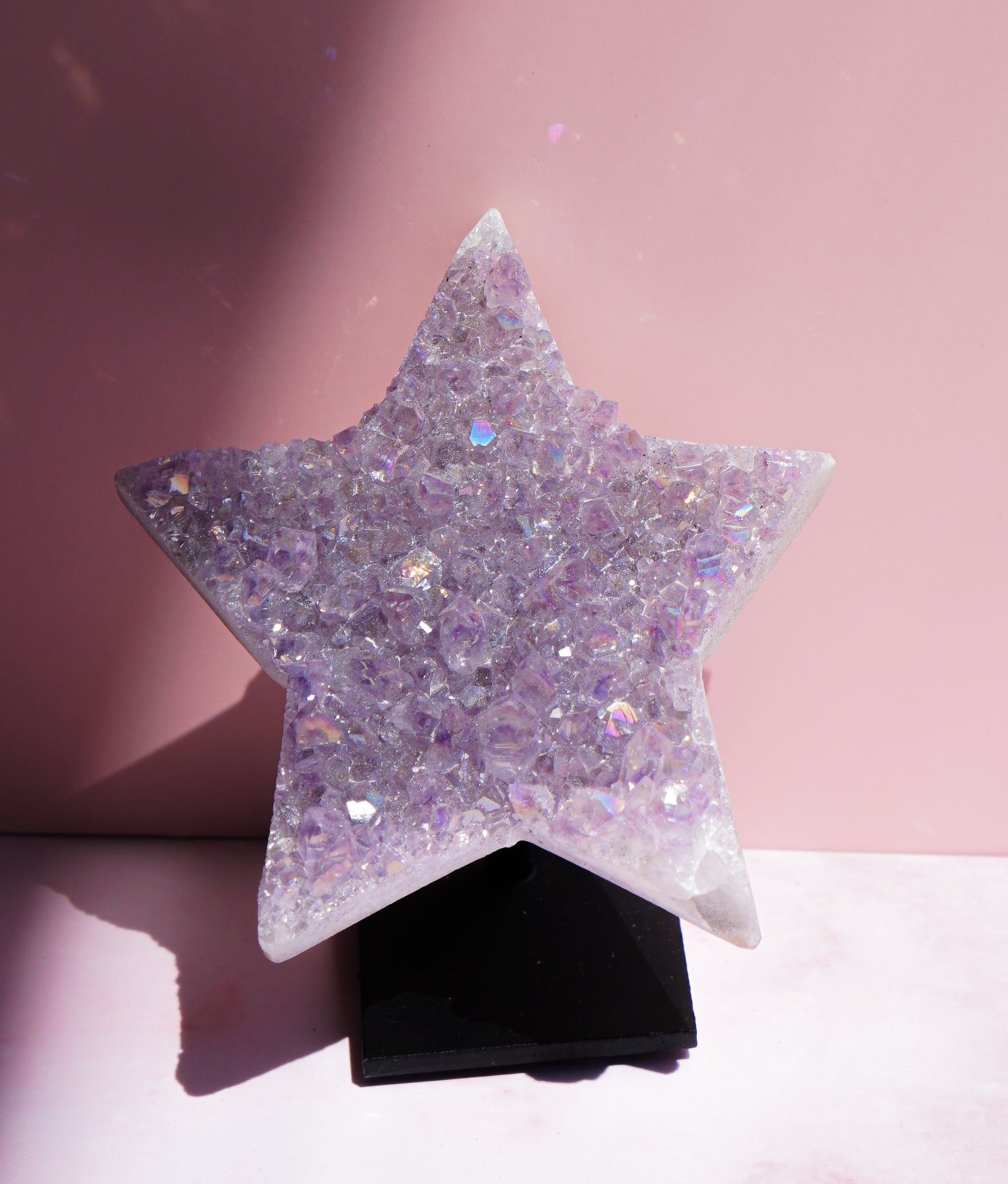 A five-pointed star-shaped Aura Amethyst raw crystal carved cluster with iridescent lavender and purple hues, displayed on a black stand against a pink background.