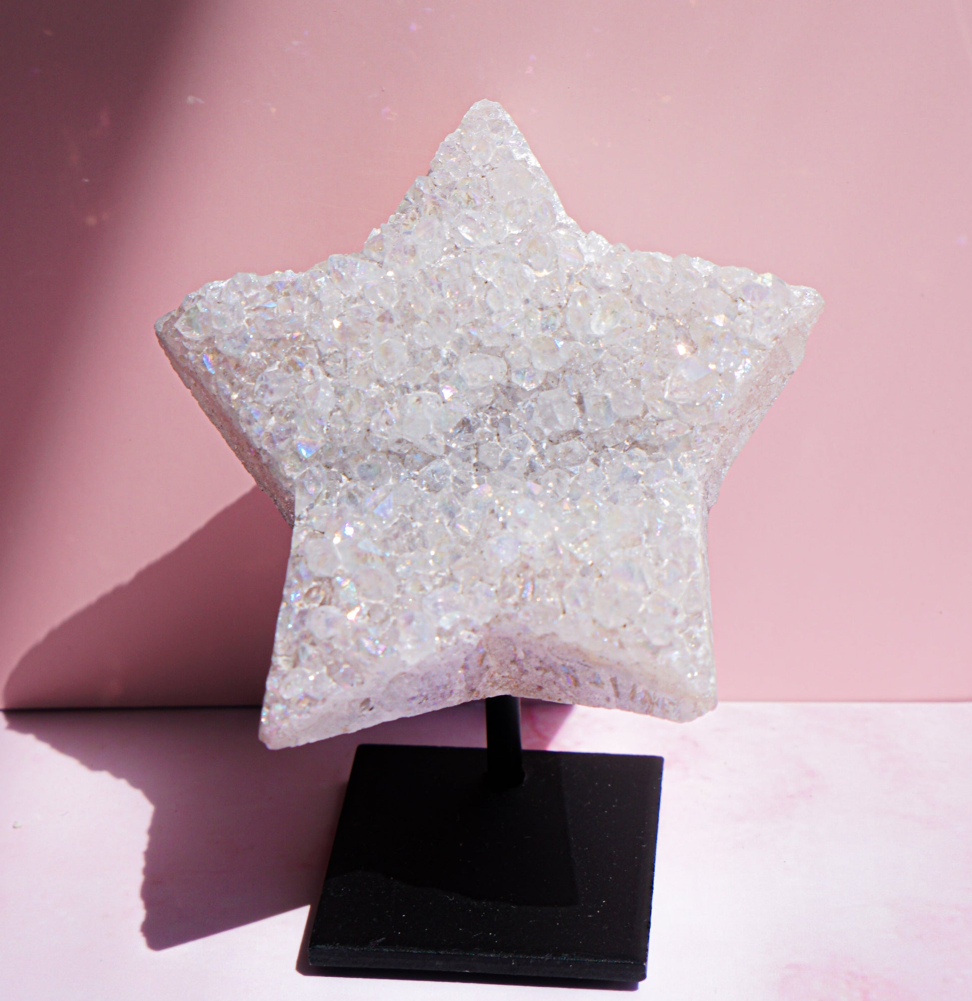 A faceted, glittery white quartz aura star-shaped raw crystal or decorative object displayed on a black stand against a pink studio background.