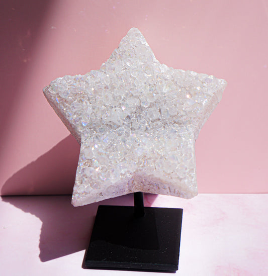 A faceted, glittery white quartz aura star-shaped raw crystal or decorative object displayed on a black stand against a pink studio background.