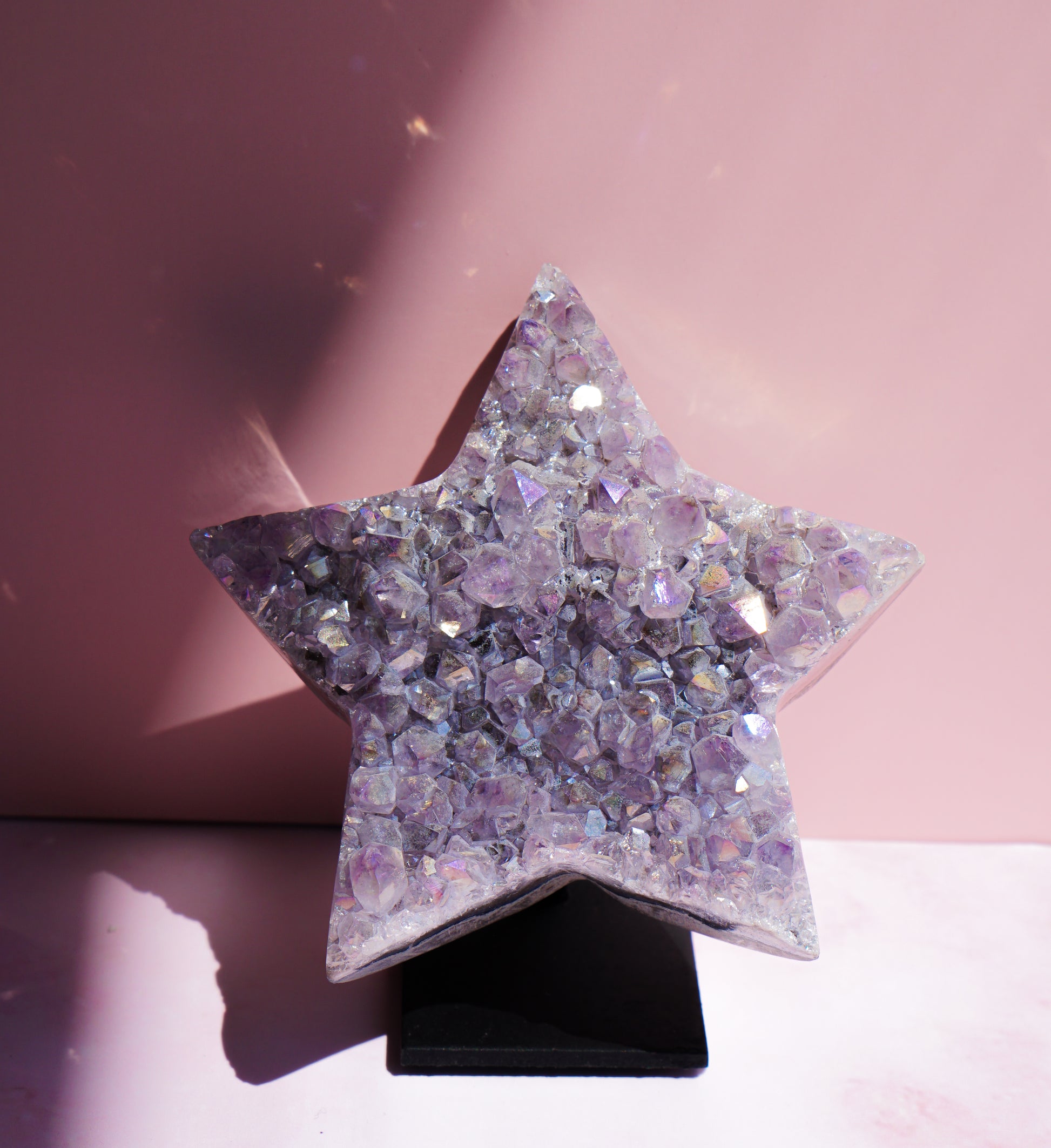 A five-pointed star-shaped Aura Amethyst raw crystal carved cluster with iridescent lavender and purple hues, displayed on a black stand against a pink background.