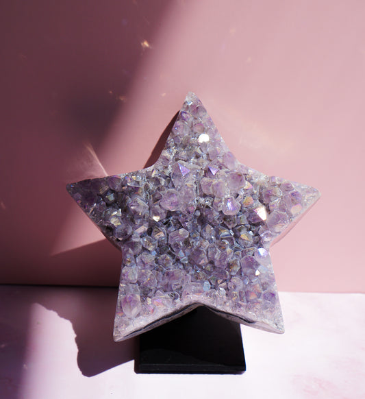 A five-pointed star-shaped Aura Amethyst raw crystal carved cluster with iridescent lavender and purple hues, displayed on a black stand against a pink background.