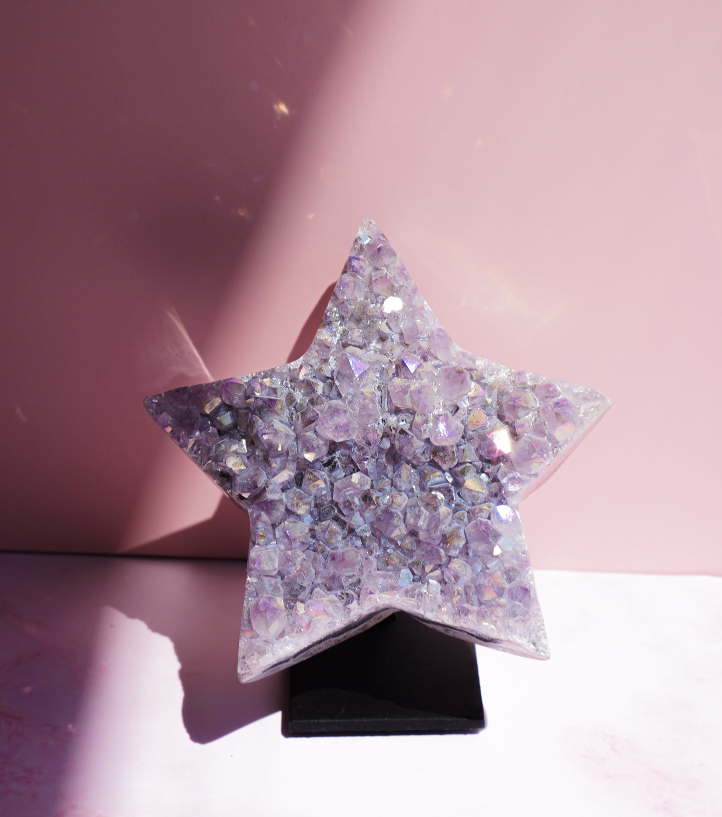 A five-pointed star-shaped Aura Amethyst raw crystal carved cluster with iridescent lavender and purple hues, displayed on a black stand against a pink background.