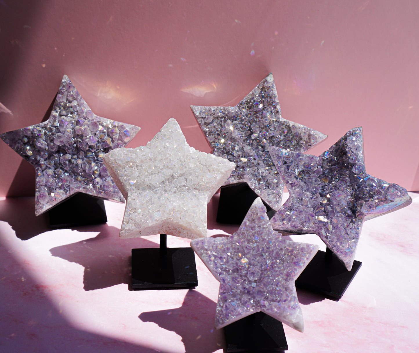 Five star-shaped crystal geode ornaments with iridescent glitter and druzy stone, displayed on black stands against a pink background.