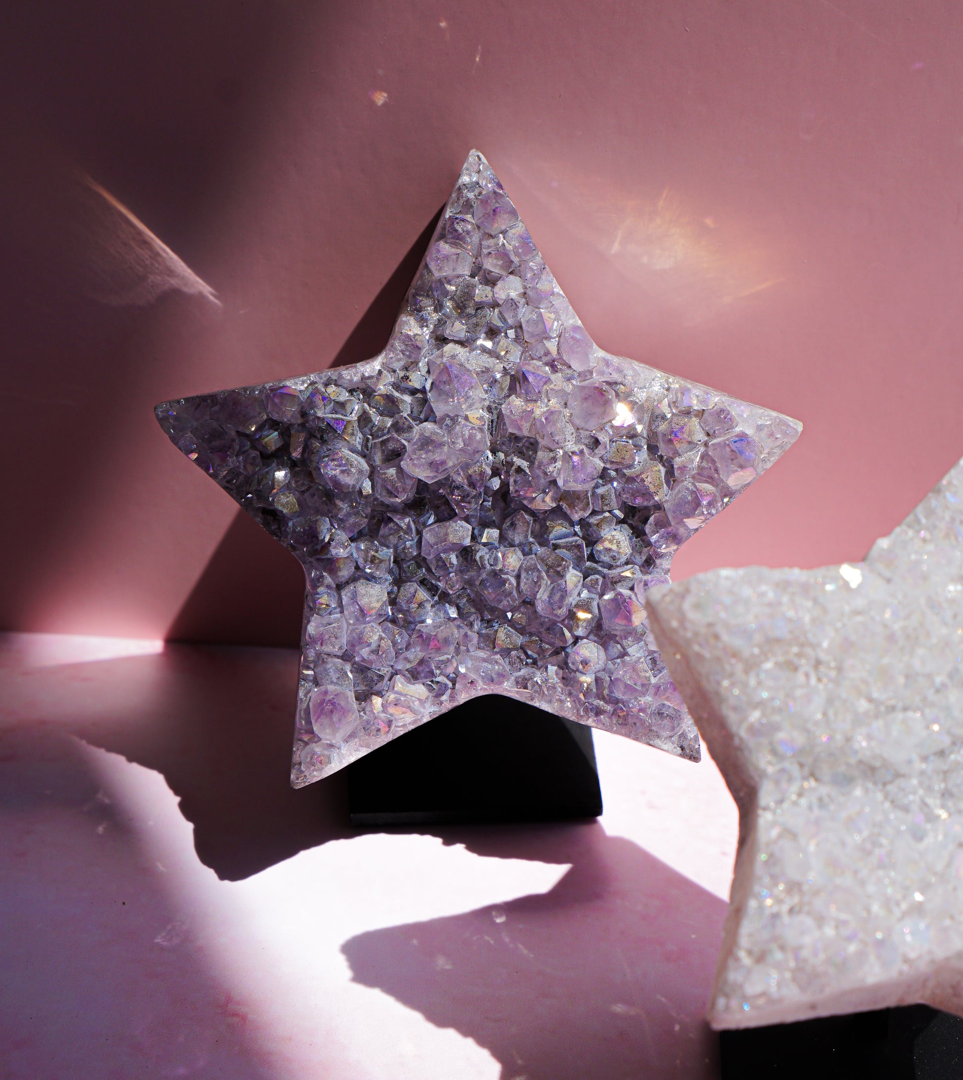A star-shaped crystal cluster ornament with faceted iridescent purple and pink stones on a black stand, displayed against a pink background with another crystal star partially visible in the foreground.