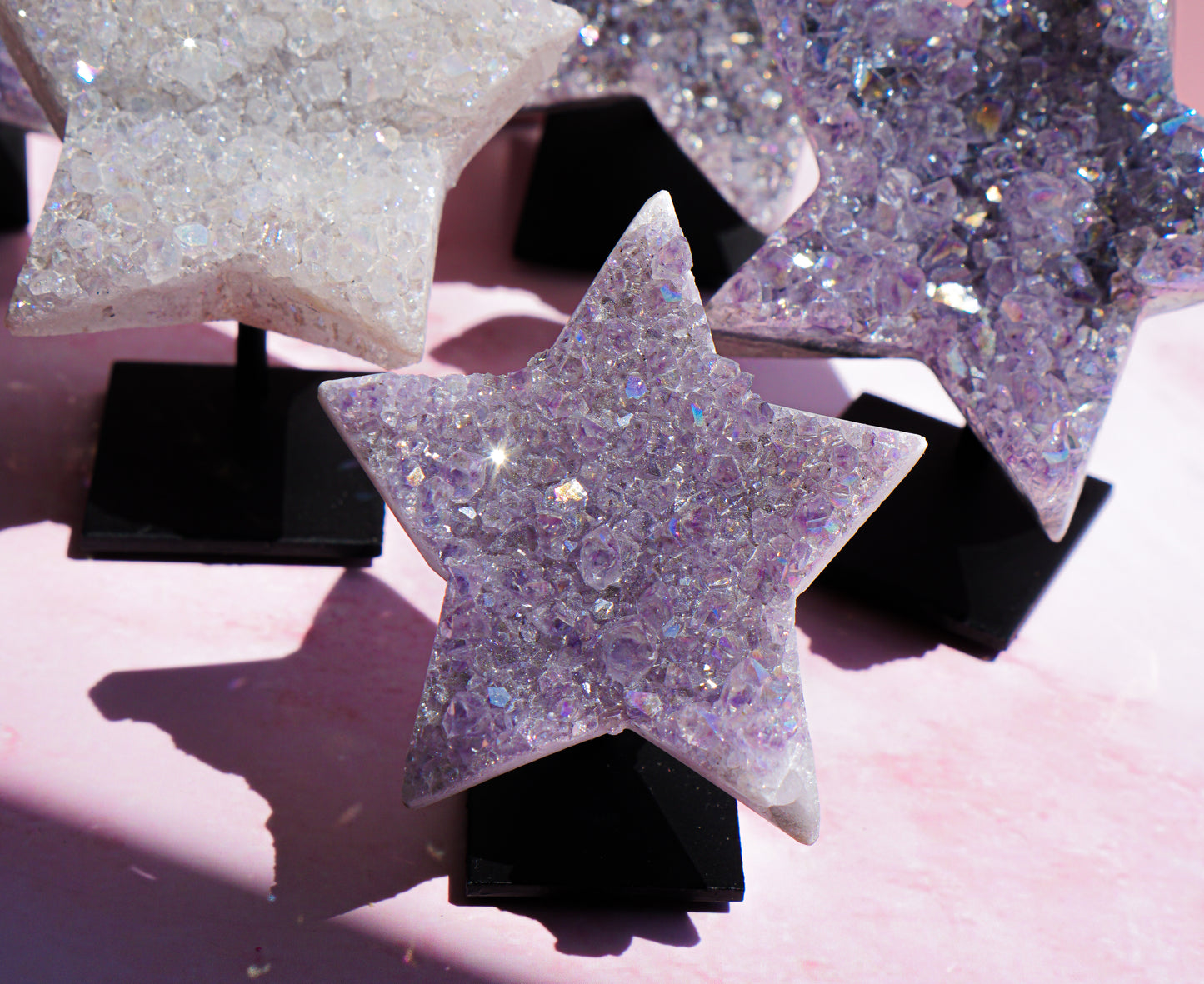 Three star-shaped crystal clusters on black display stands, featuring lavender amethyst and clear quartz with iridescent glitter, set against a soft pink background.