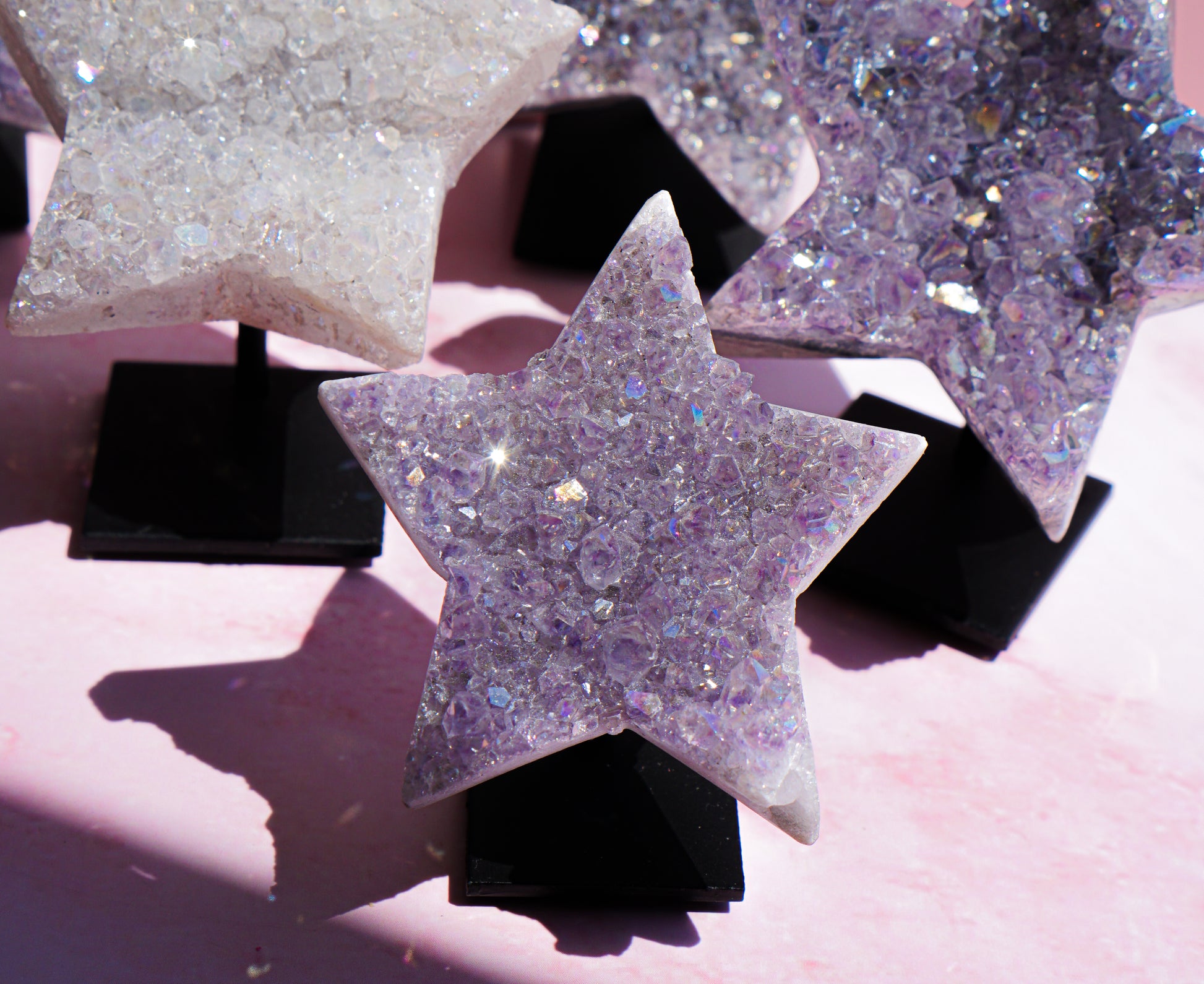Three star-shaped crystal clusters on black display stands, featuring lavender amethyst and clear quartz with iridescent glitter, set against a soft pink background.