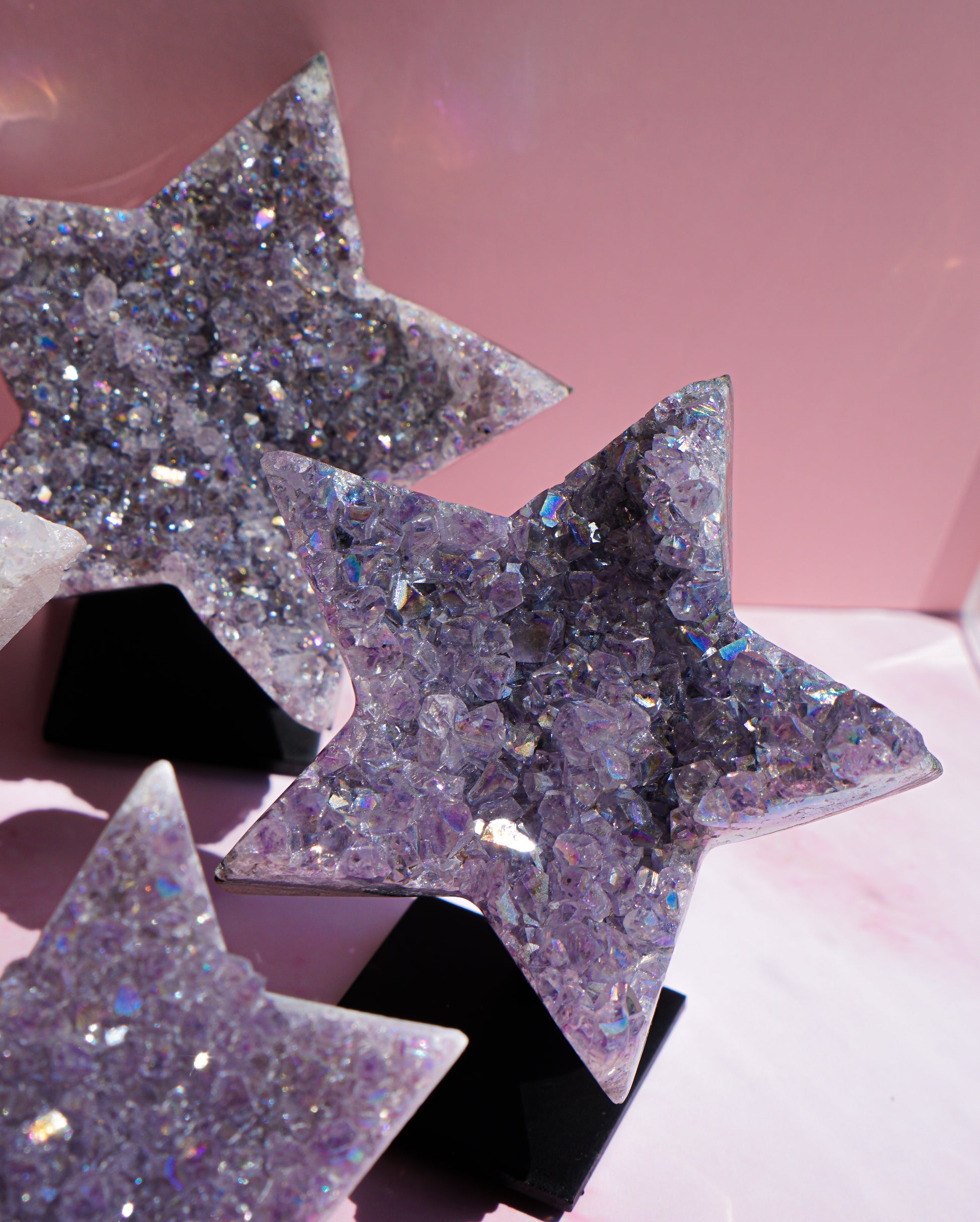 Three star-shaped resin ornaments filled with sparkling lavender and silver glitter, displayed on black stands against a soft pink background.