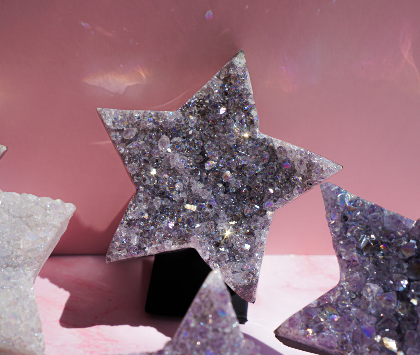 A glittery star-shaped crystal cluster ornament in lavender and silver, displayed on a pink surface with other star ornaments partially visible.