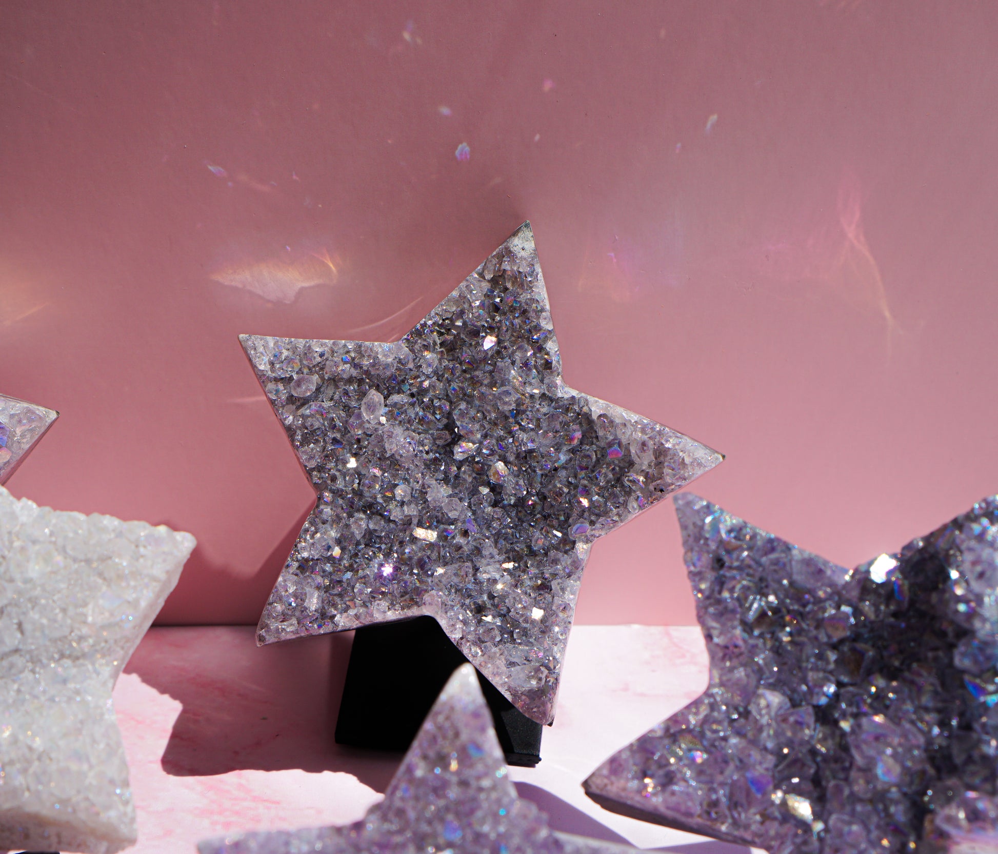 A glittery star-shaped crystal ornament in lavender and purple tones, displayed on a black stand against a pink background with other star ornaments partially visible.
