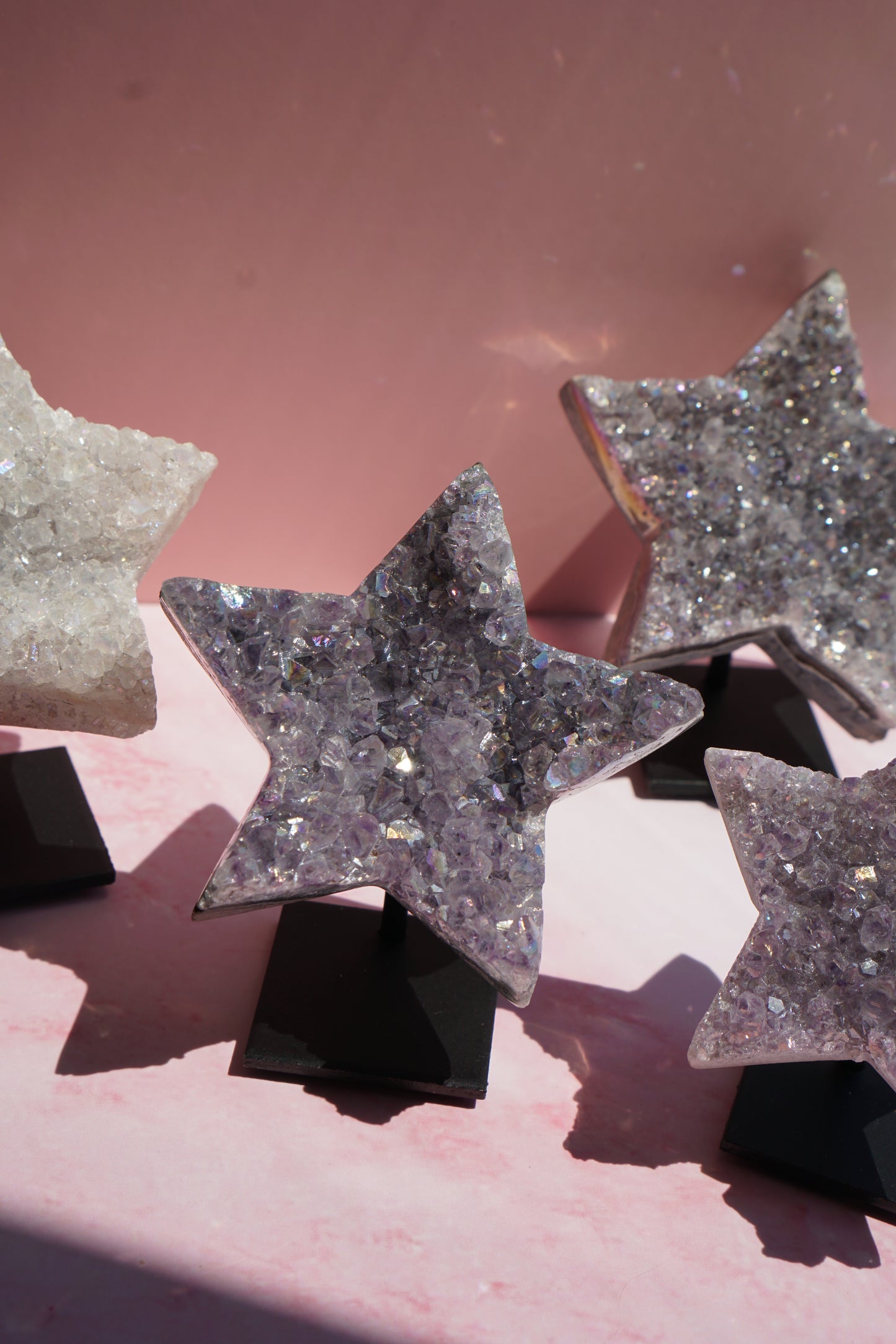 Three star-shaped crystal druzy ornaments on black display stands, set against a pink background with dramatic shadows.
