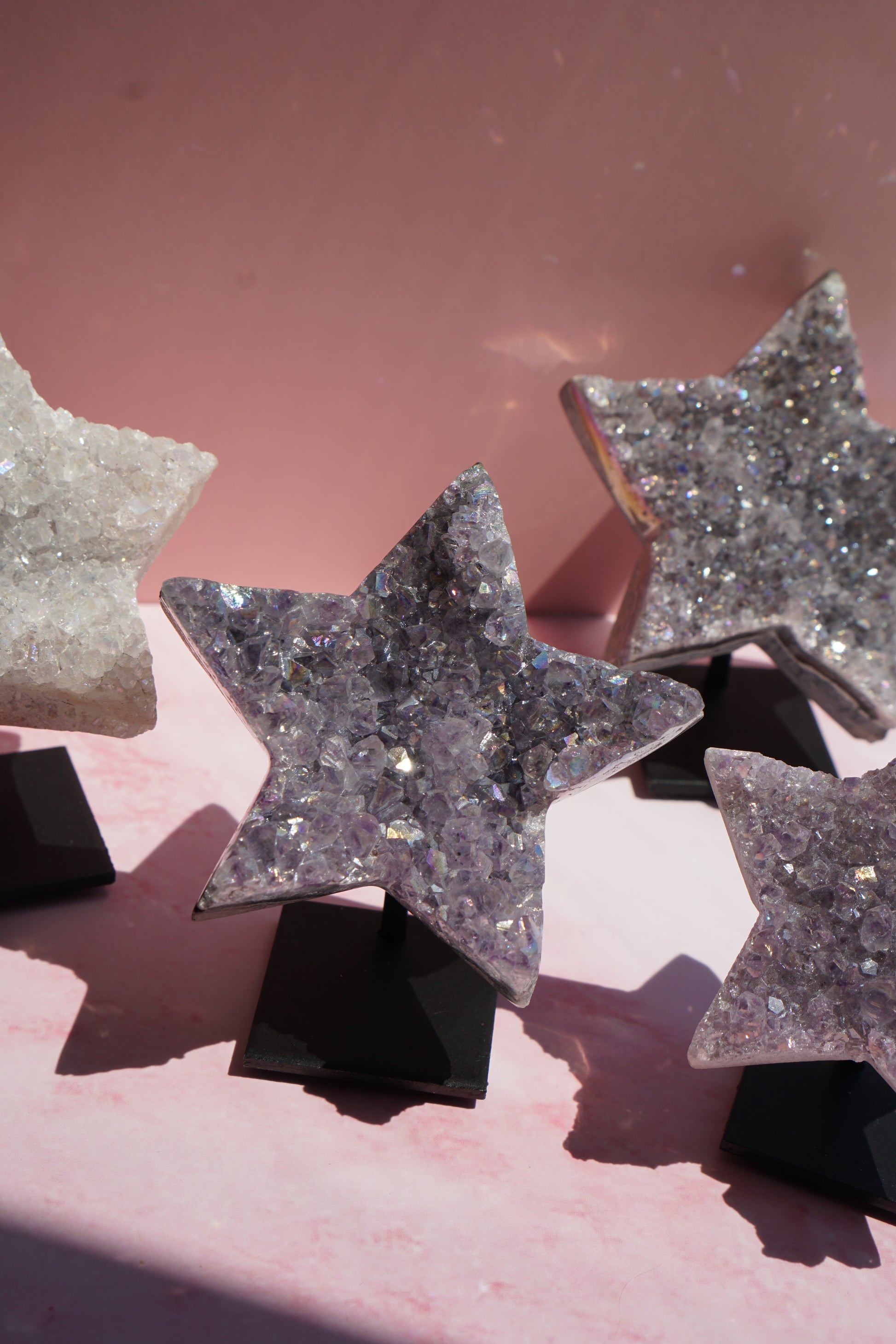 Three star-shaped crystal druzy ornaments on black display stands, set against a pink background with dramatic shadows.