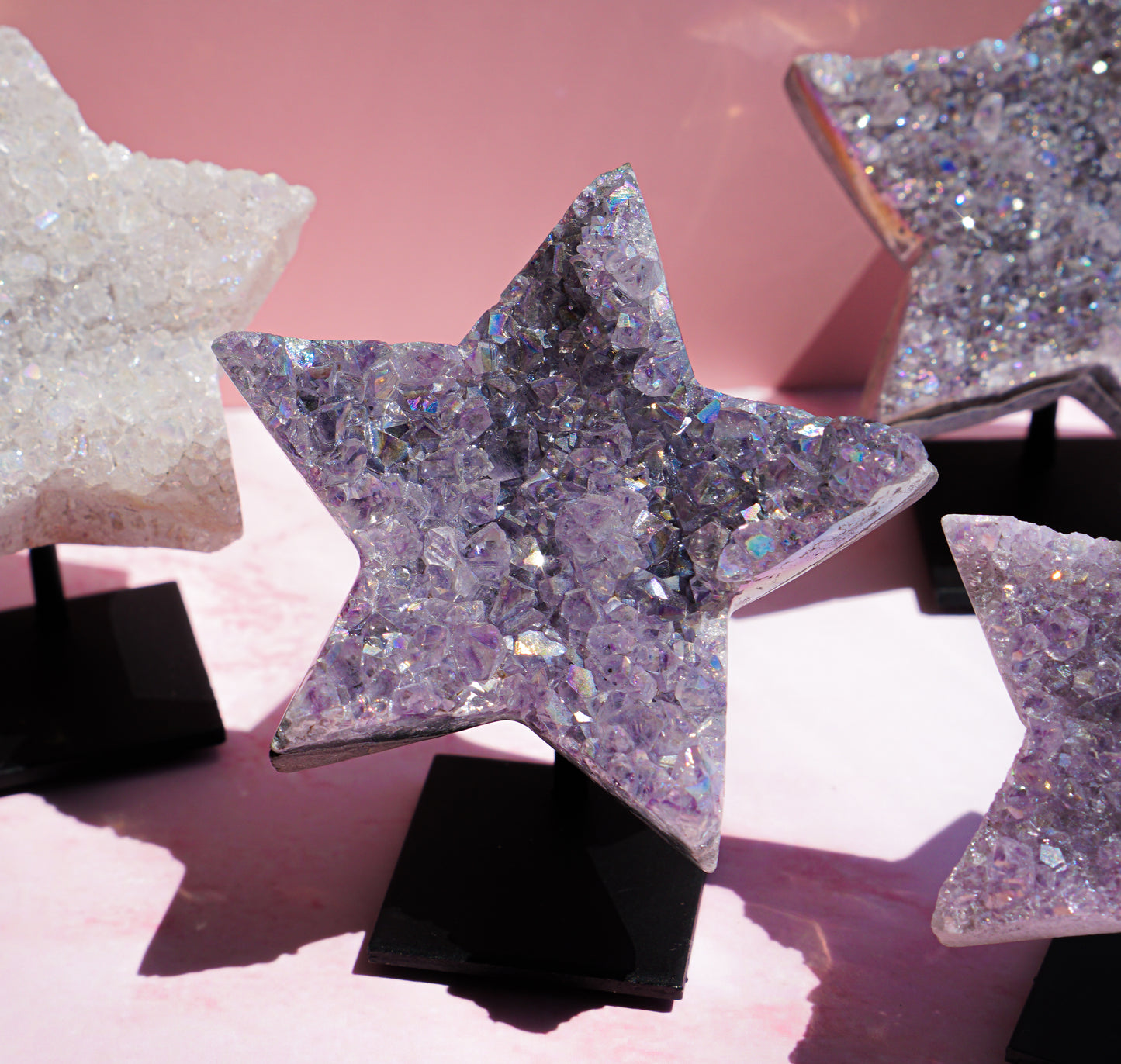 Star-shaped crystal clusters with iridescent glitter, displayed on black stands against a soft pink background.