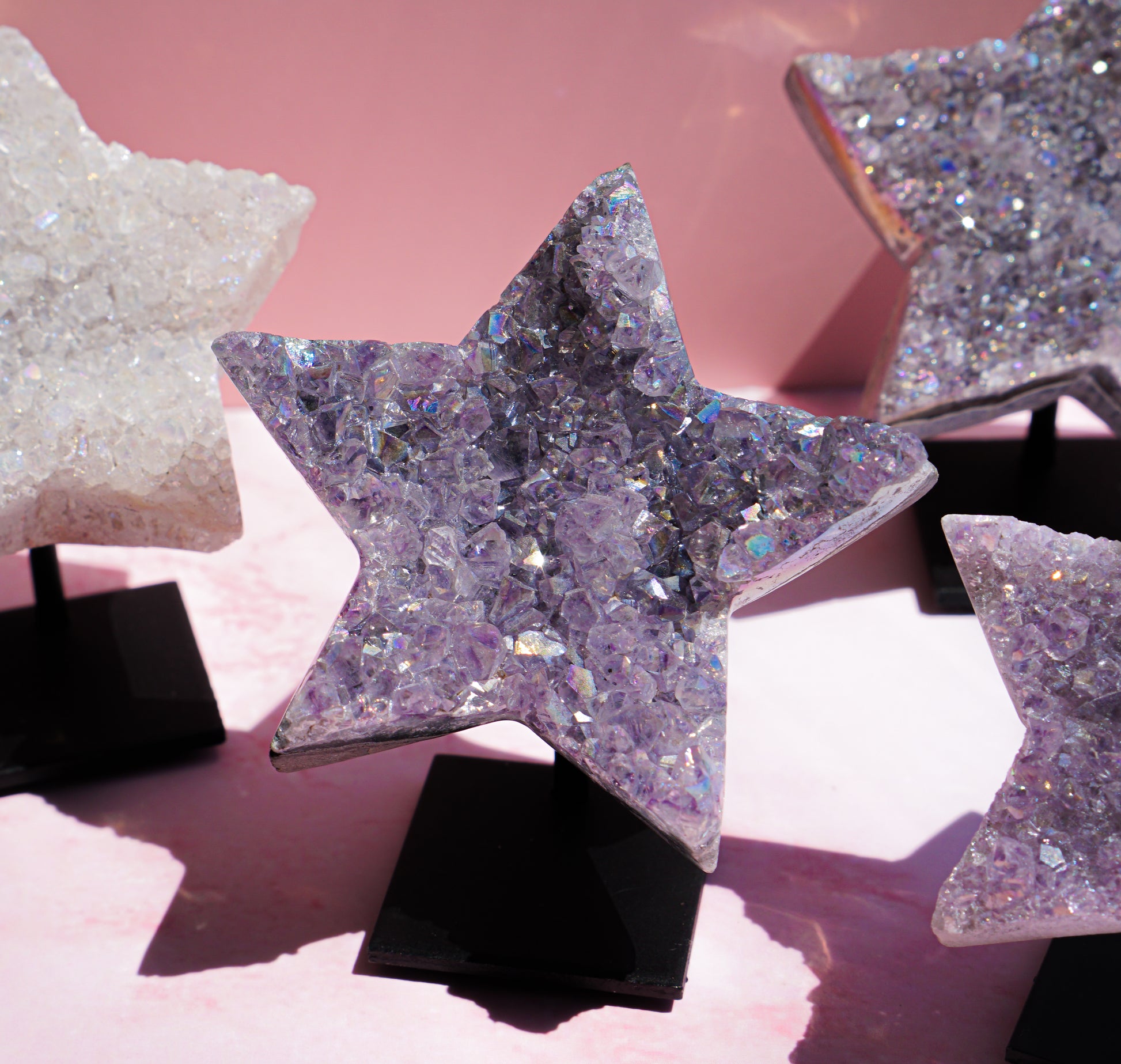 Star-shaped crystal clusters with iridescent glitter, displayed on black stands against a soft pink background.